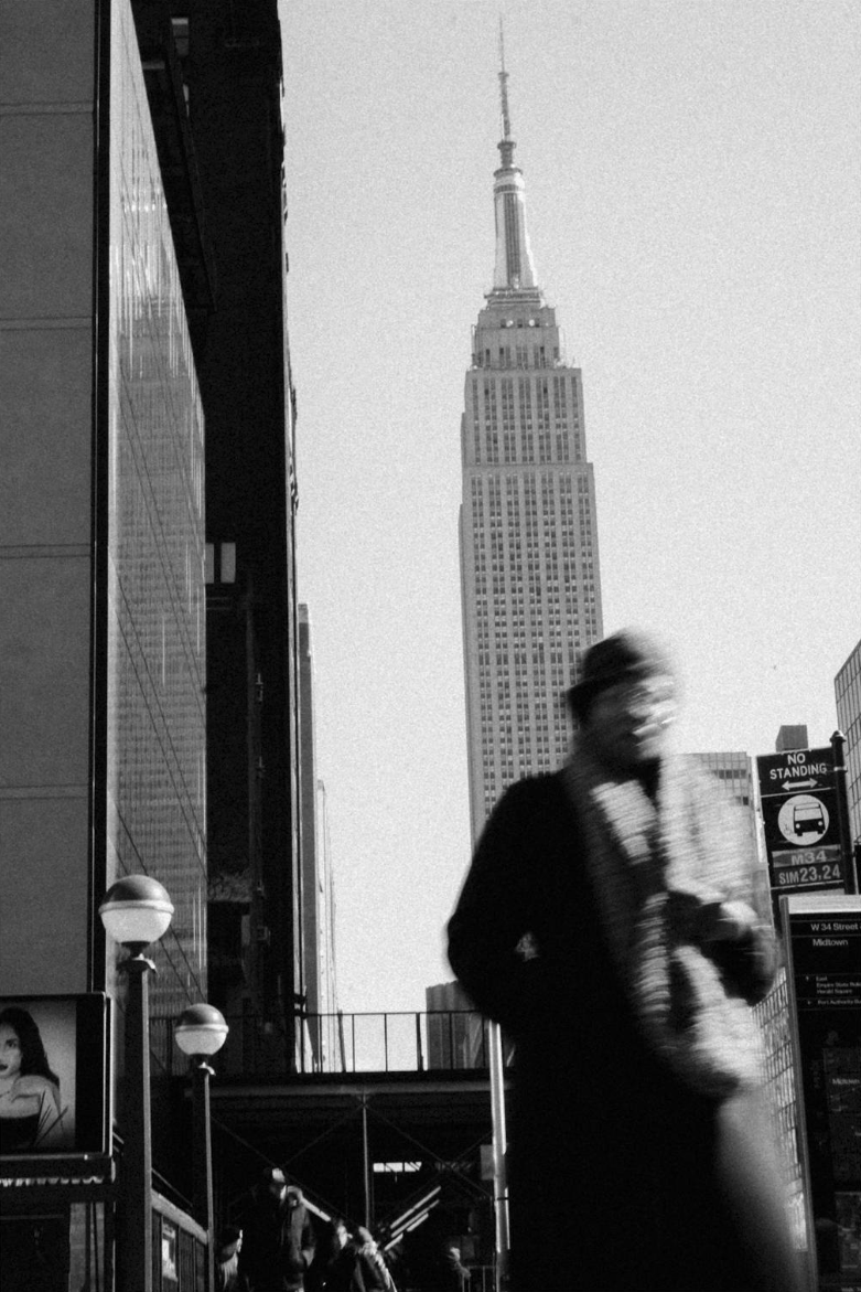 Black and white photo of the Empire State Building in New York City with a blurred person in the foreground walking past the street.