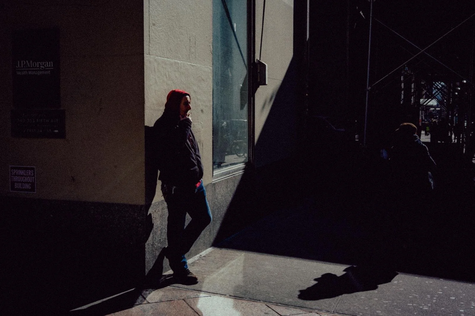 A person wearing a red hat and black jacket leaning against a building, with their hand near their face, in shadow on a city sidewalk.