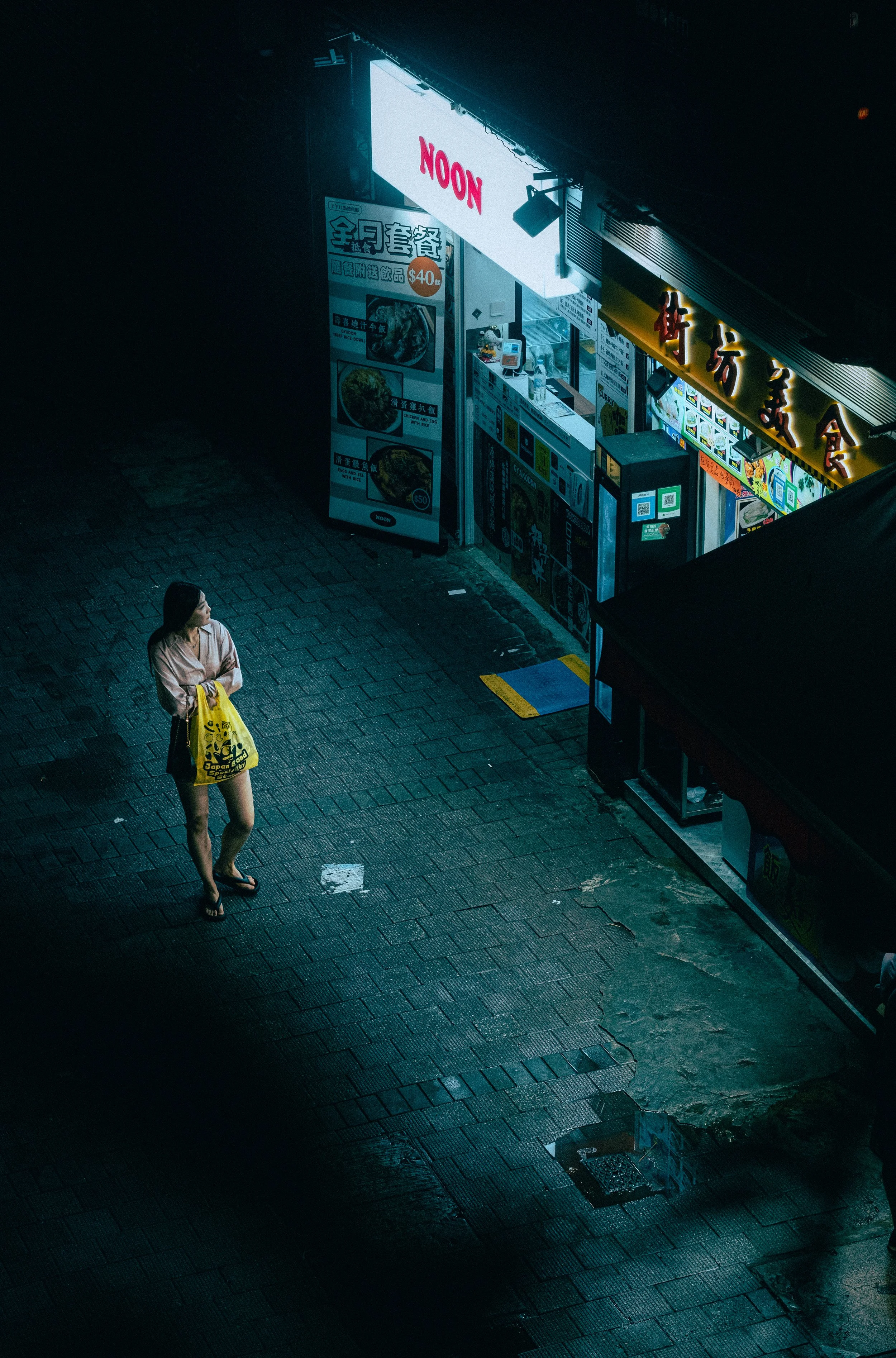 A woman standing outside a brightly lit restaurant at night, holding a yellow shopping bag, looking at the menu and neon signs in an urban setting.