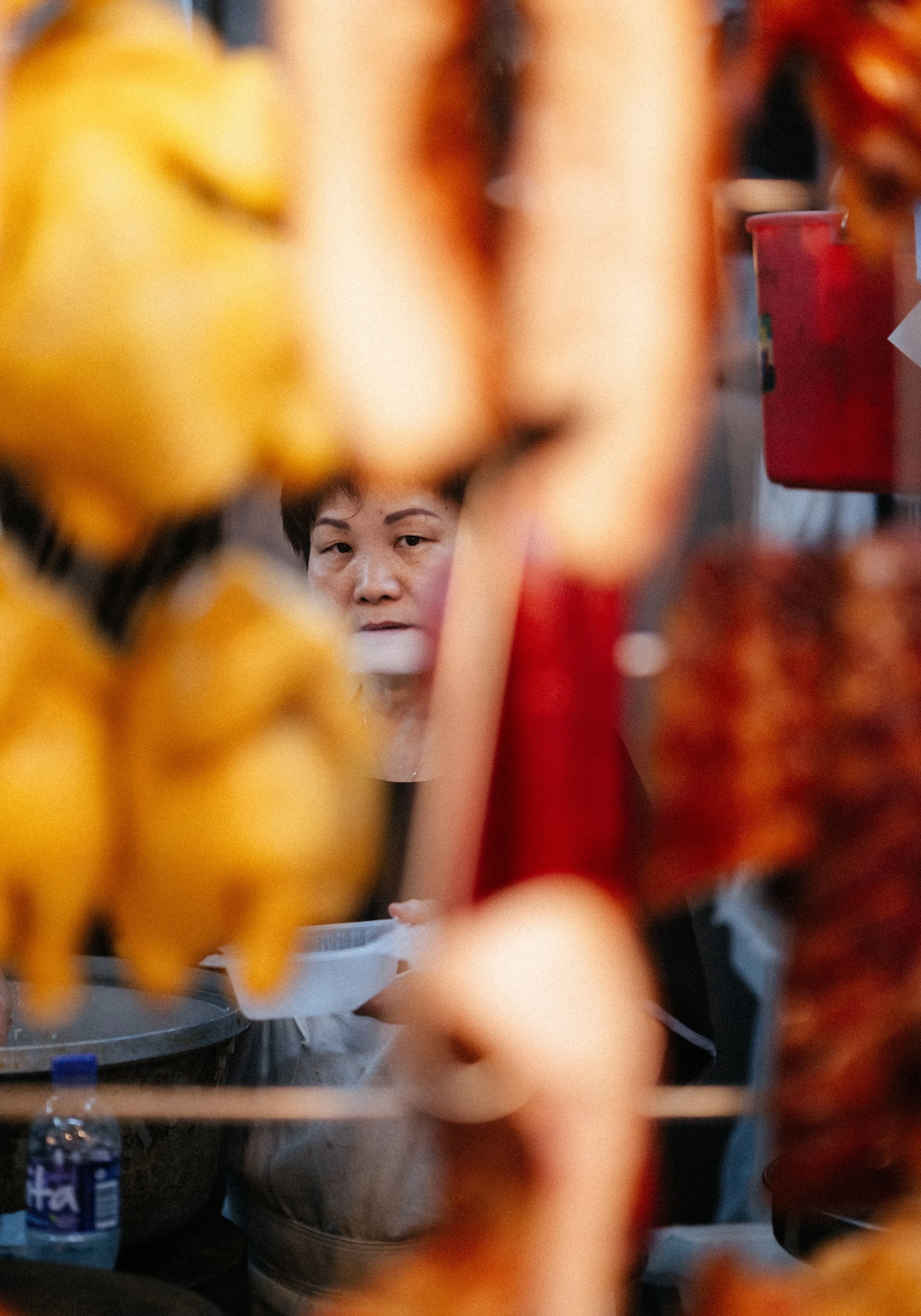 A woman is looking through a gap at a street food stall with skewers of yellow and red grilled food hanging in the foreground.