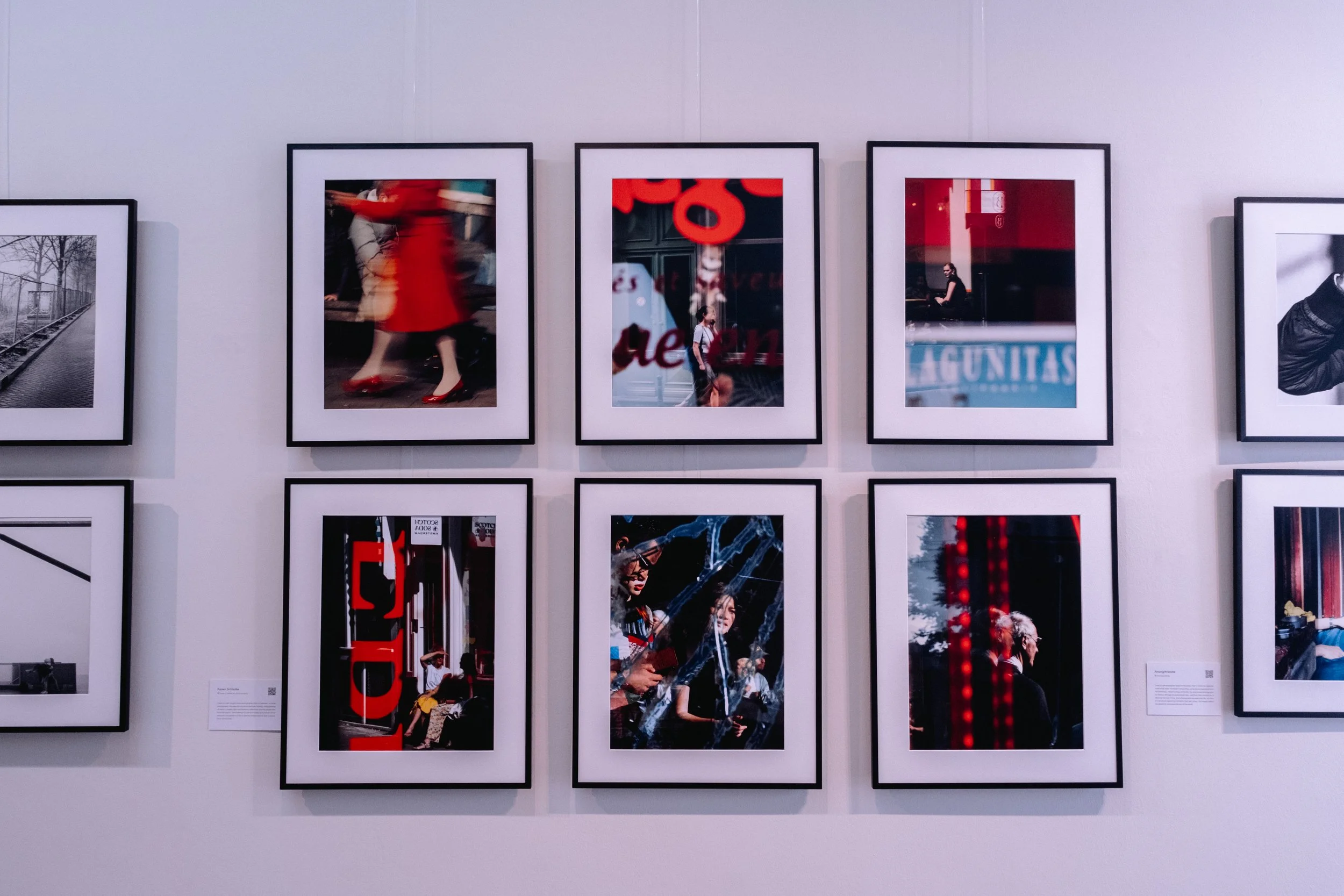Street Photography art gallery in Amsterdam with framed photographs displayed on white walls, featuring silhouettes, reflections, and urban scenes, with a partly visible art desk and chairs in background.