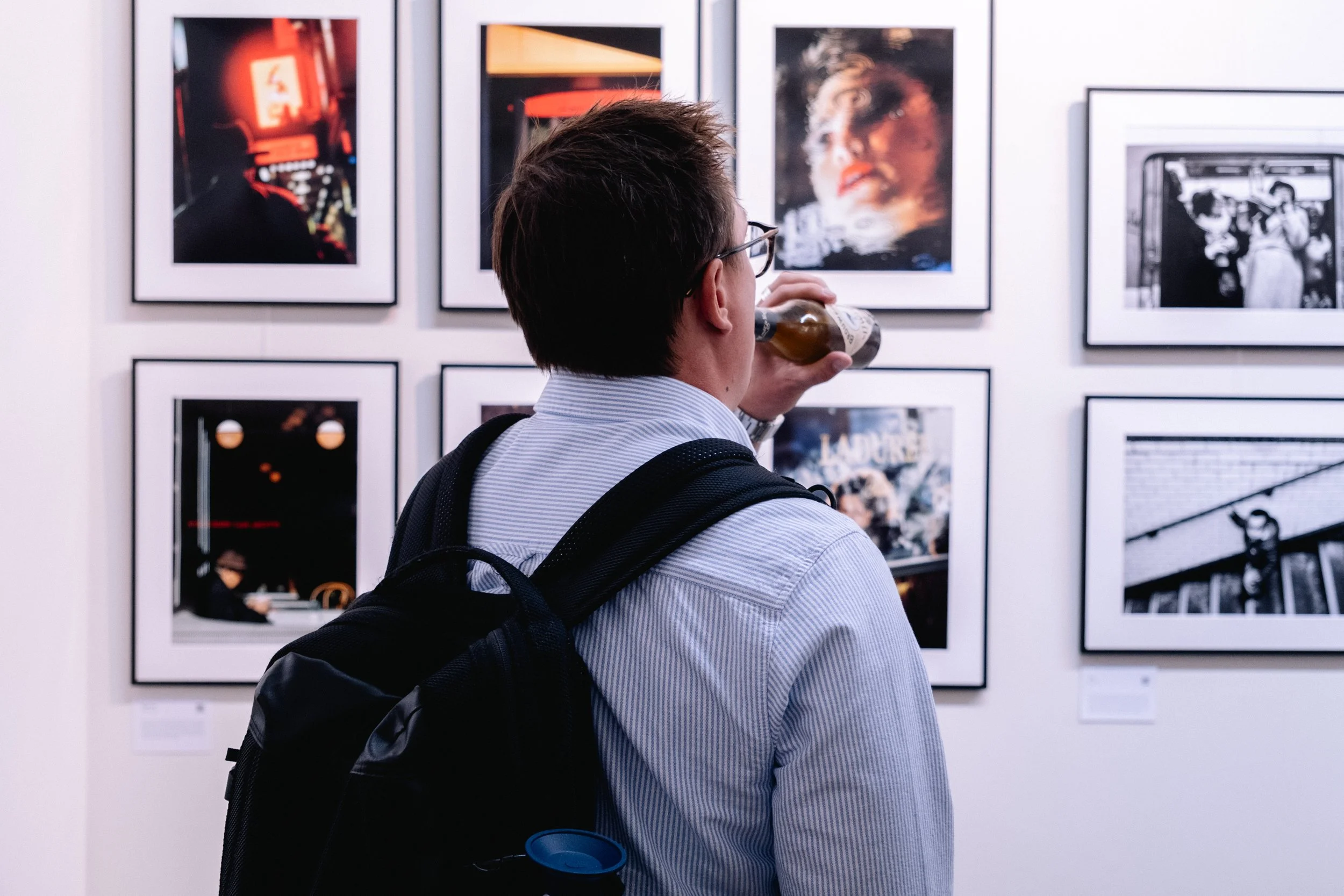 A man with glasses and a backpack looking at framed photographs on a white gallery wall while drinking from a bottle.