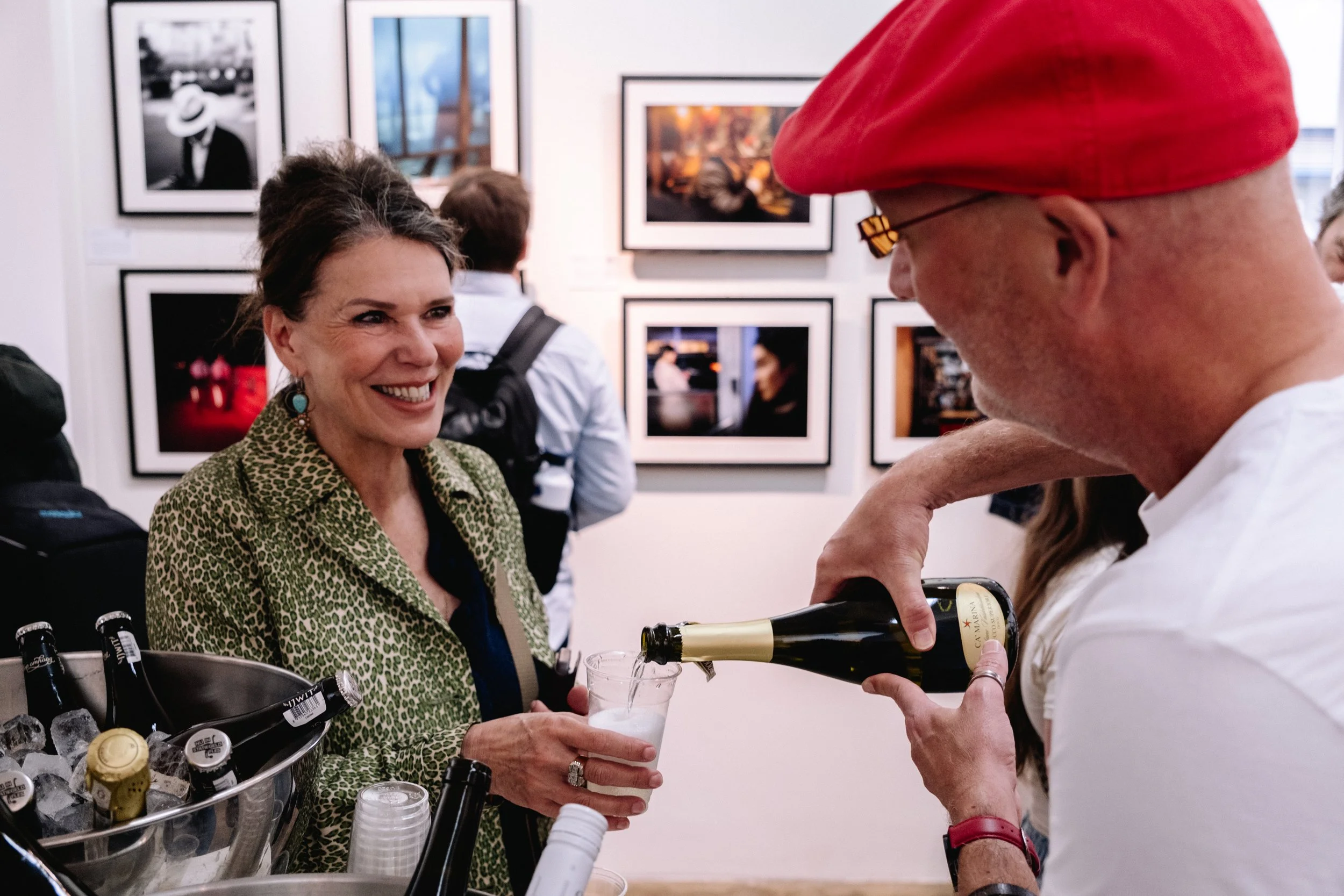 A woman smiling as a man pours champagne into her glass at an art gallery event.