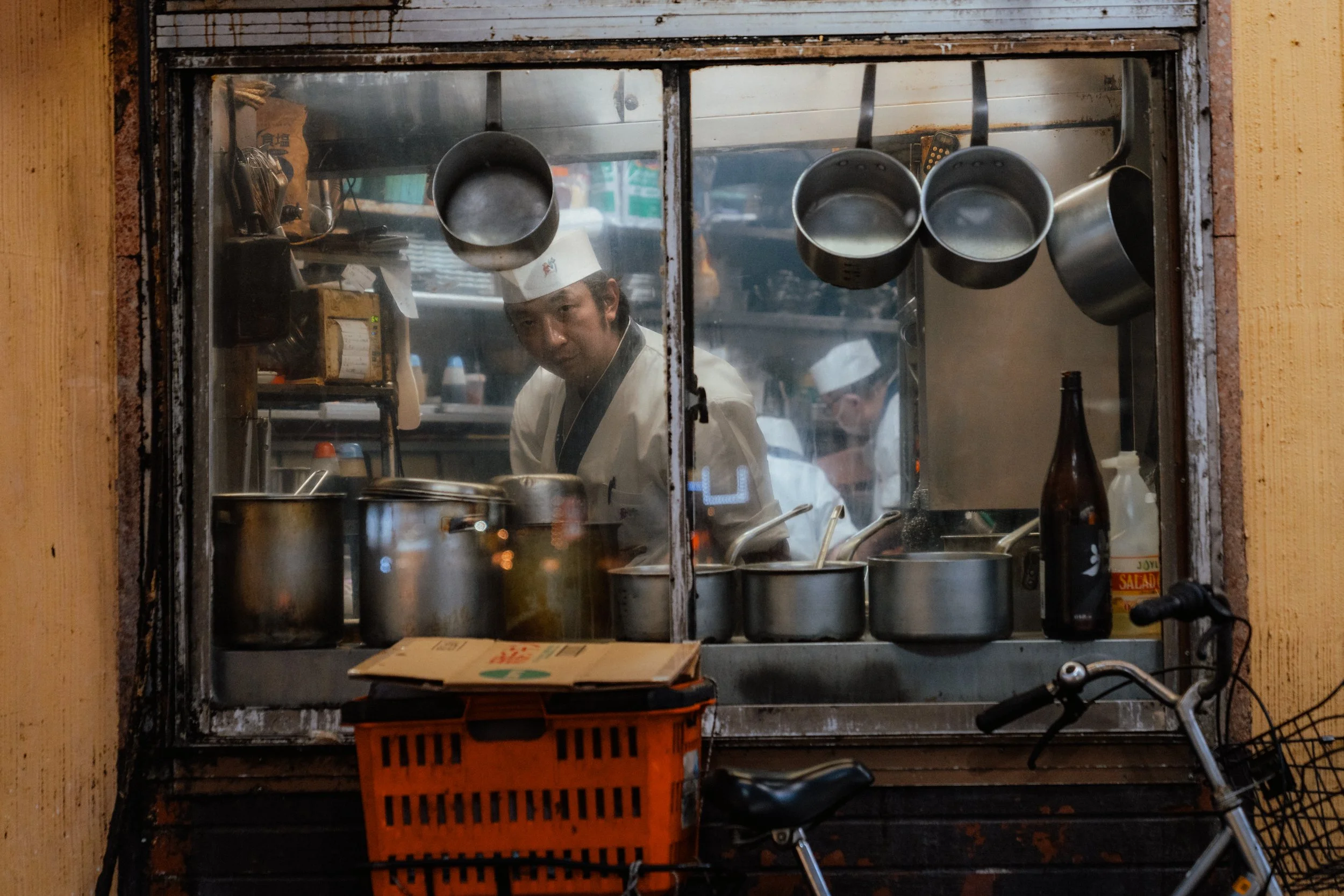 View of a busy restaurant kitchen through a window, with two chefs wearing white uniforms and hats, cooking and preparing food. Pots and utensils hang on the wall inside.