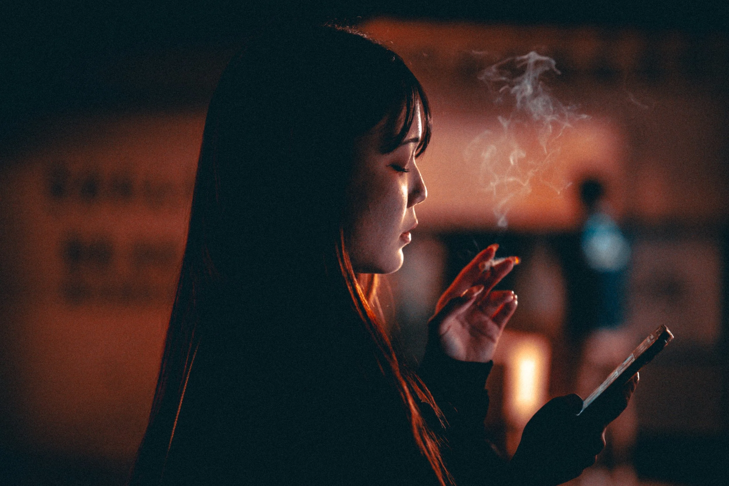 A woman with straight, dark hair in profile, smoking and looking at her smartphone in a dimly lit, warm-toned environment with blurred background