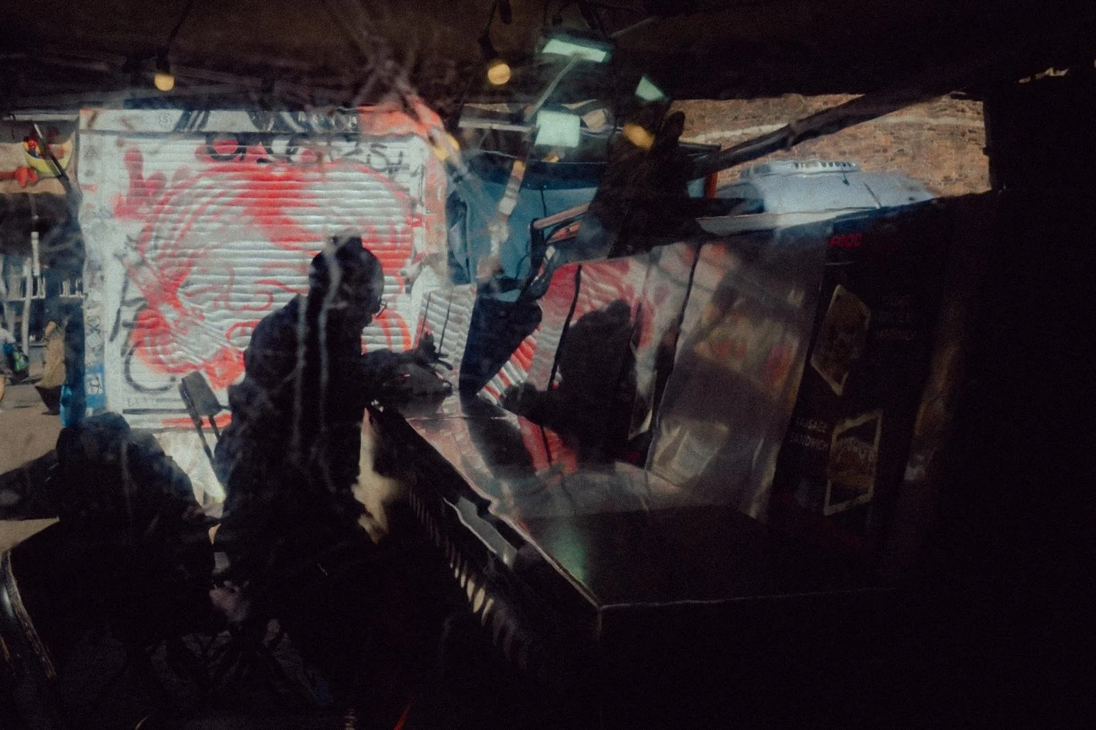 View through a dirt-streaked windshield showing a black and white dog sitting inside a vehicle with a cluttered interior, reflecting the back of the driver and the outside scene with a large red and white graffiti-style mural on a wall.
