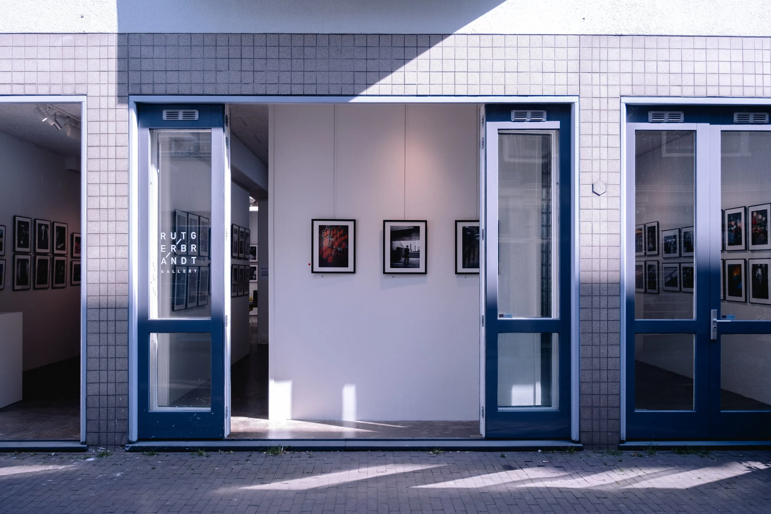 Entrance to an art gallery with open glass doors, white walls displaying framed photographs, and a glimpse of interior artwork.