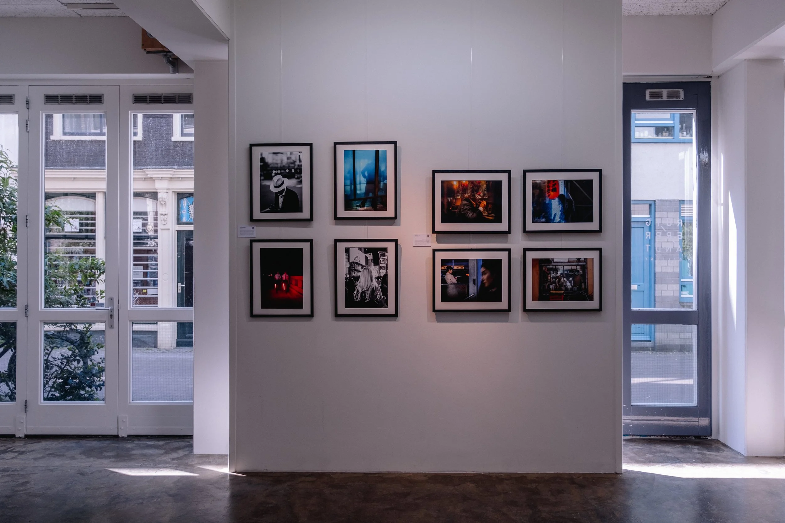 An art gallery with framed photographs displayed on a white wall, illuminated by natural light from large windows on either side.