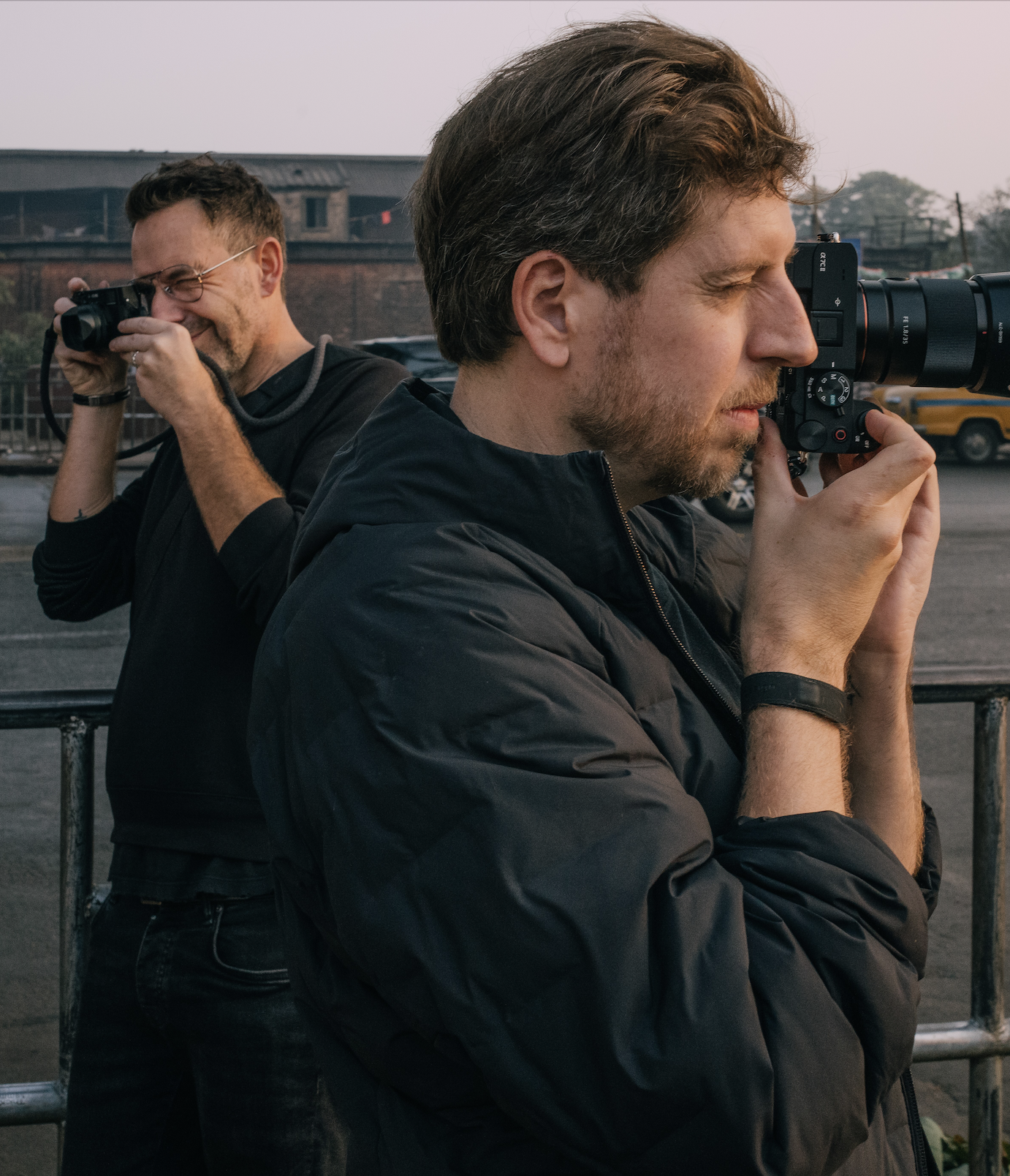 Two men are taking photographs outdoors during dusk. The man in the foreground has light brown hair, a beard, and is using a camera with a large lens. The man in the background has short hair, glasses, and is smiling while looking through the viewfinder of his camera.