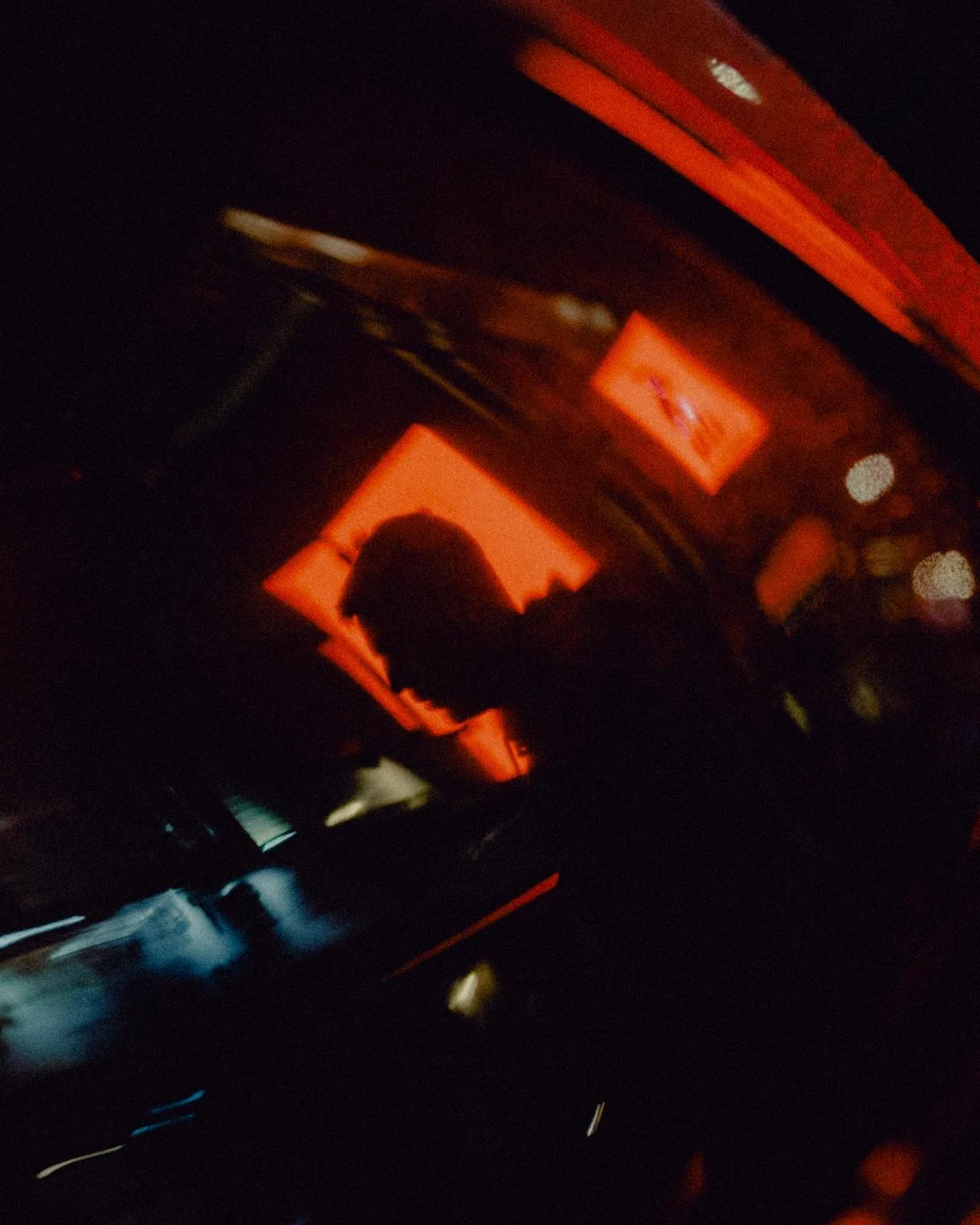 A person with glasses and short hair in silhouette is reflected in a car window, with red interior lighting and moving light streaks in the background.