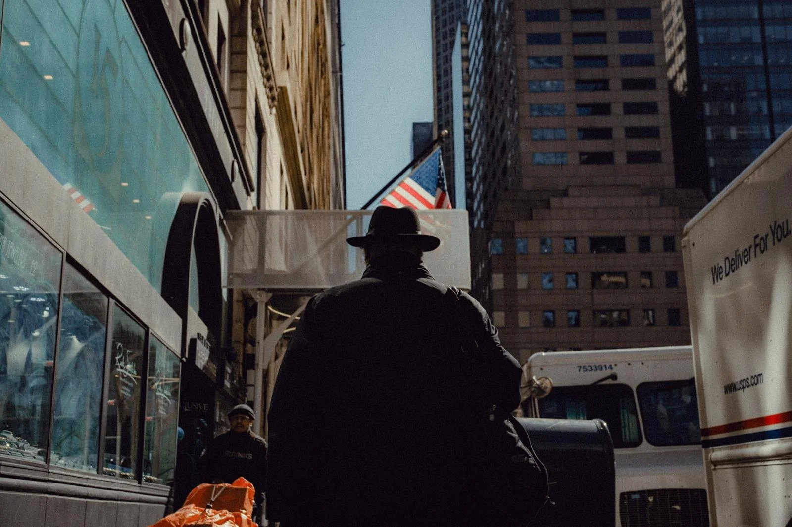 A man wearing a black coat and wide-brimmed hat is seen from behind, standing on a city street surrounded by tall buildings, with a United States flag visible overhead and delivery trucks nearby.