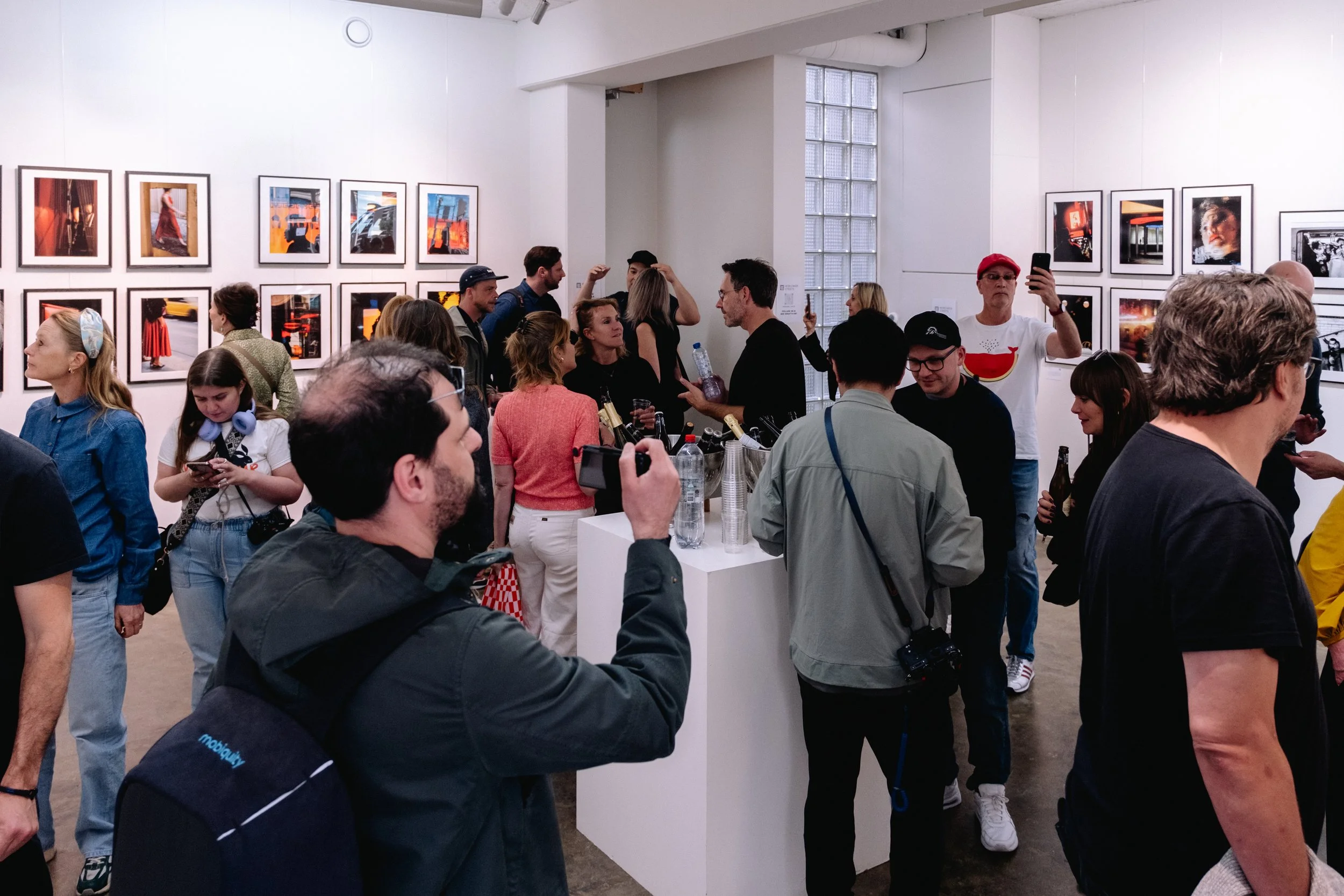 People attending an art exhibit or gallery, viewing photographs hanging on the white walls, engaging in conversations, and taking photos of the artwork.
