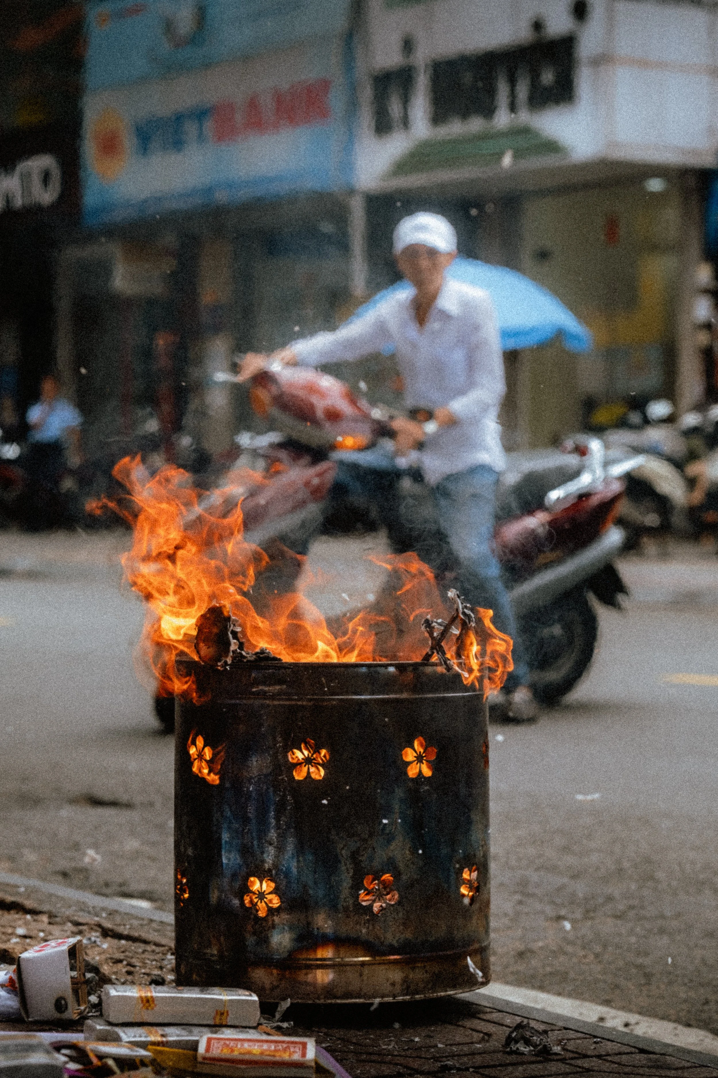 A person with a white hat and white shirt riding a scooter on a street, with a burning trash bin in the foreground and shops and other scooters in the background.