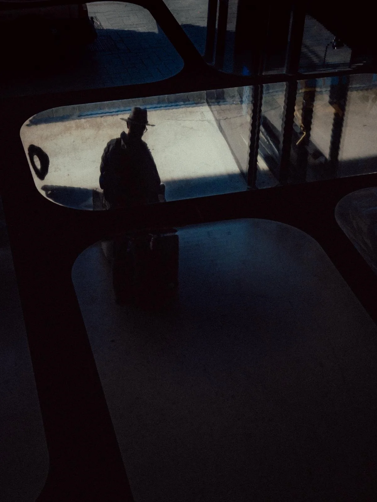 Silhouette of a man wearing a hat and backpack, reflected on a window, with a dark interior.