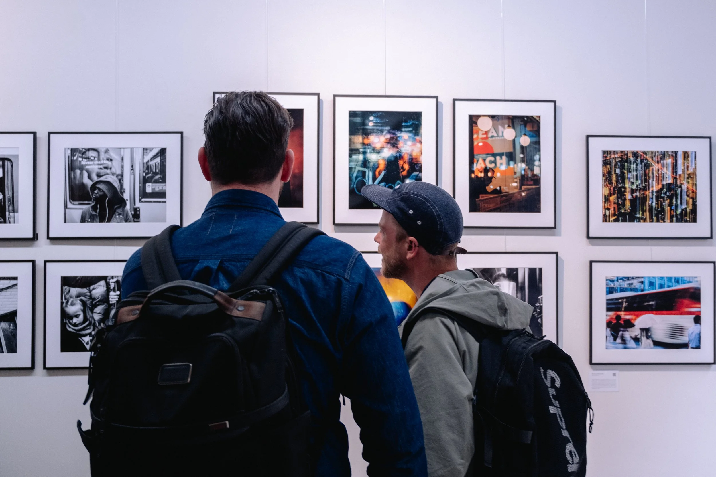 Two men with backpacks viewing framed photographs and artwork on a gallery wall.