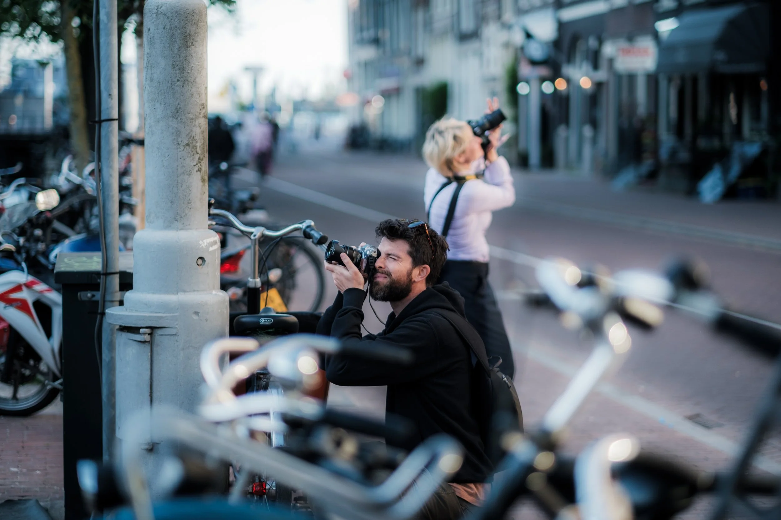 Two photographers, a man with a beard and a woman with blonde hair, are shooting photos on a city street with parked bicycles and motorcycles nearby, and buildings in the background.