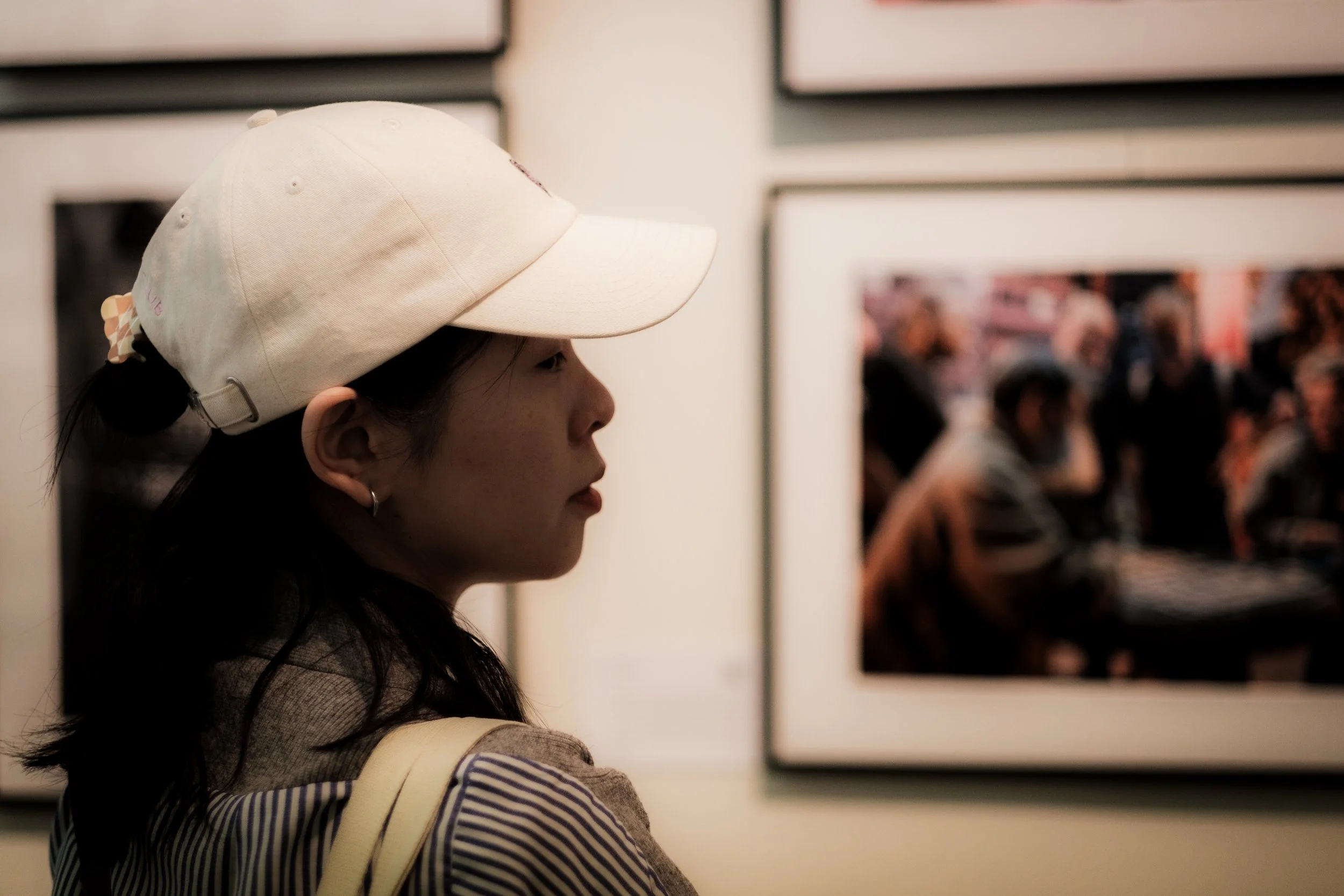 A young woman with dark hair wearing a beige baseball cap and striped shirt views framed photographs on a gallery wall.