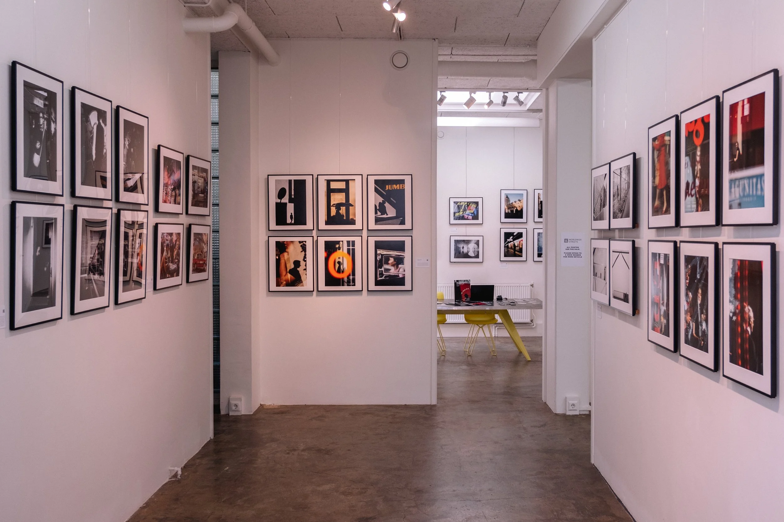 Gallery with framed photographs and artwork on white walls, wooden floor, and a workspace with a yellow chair in the back.