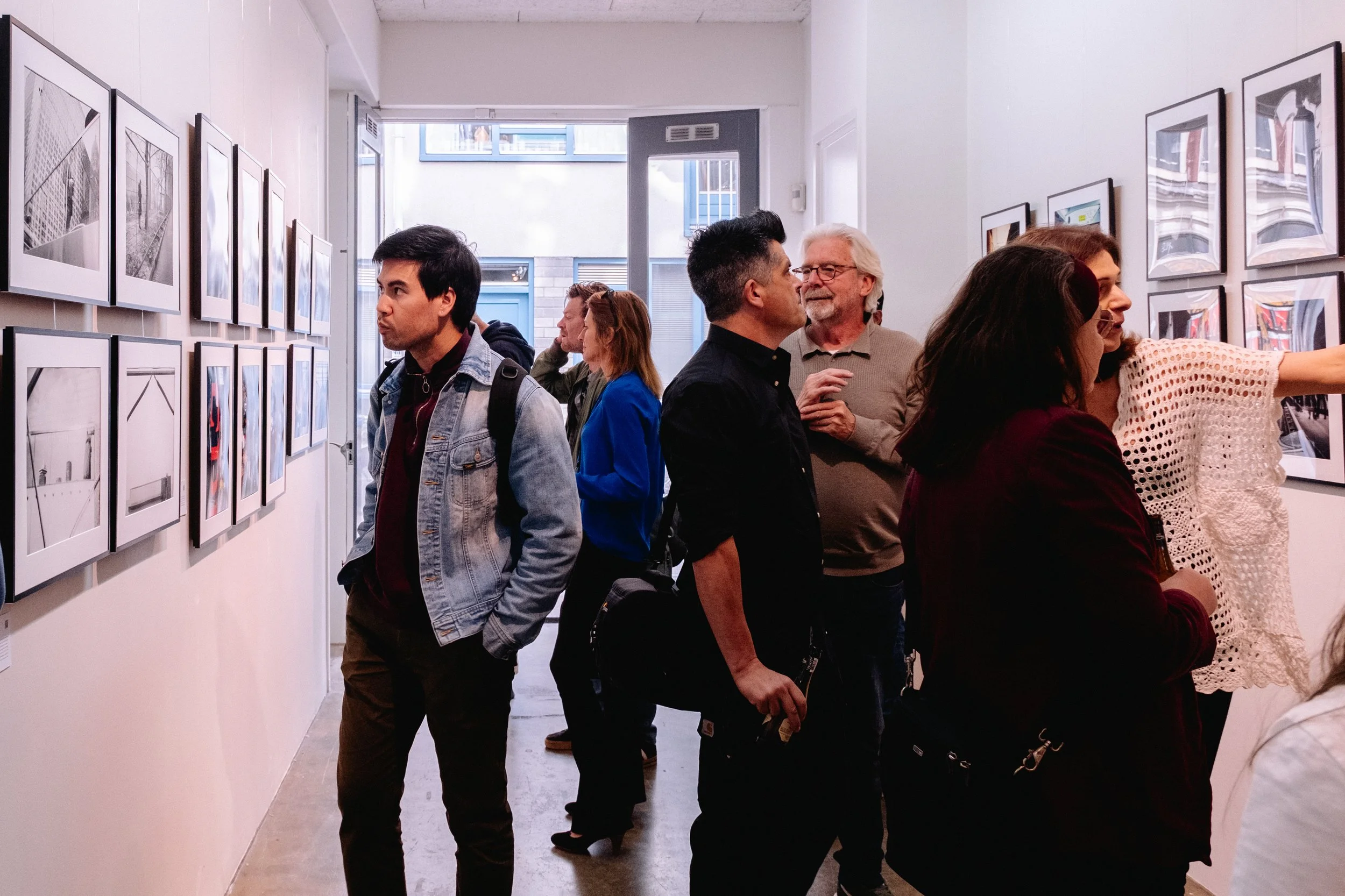 People viewing photographs displayed on white walls inside an art gallery with large windows.