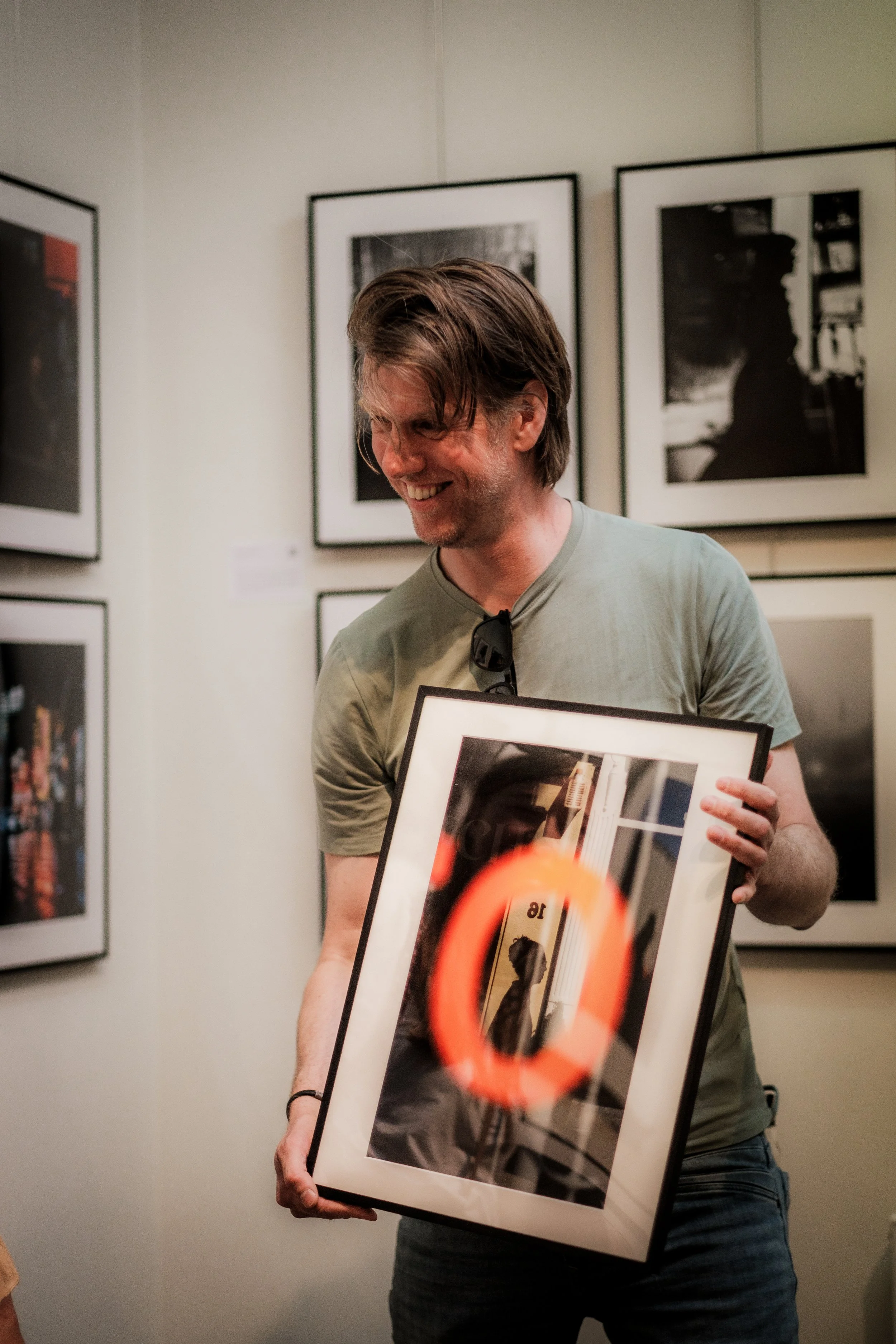 A man holding a framed photograph and smiling at an art gallery with black and white pictures on the wall behind him.