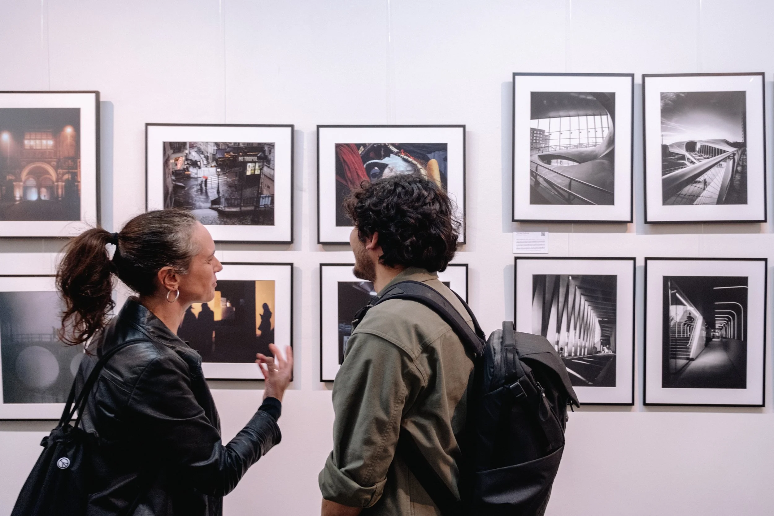 Two people are talking in an art gallery with framed black-and-white photographs on the wall behind them.