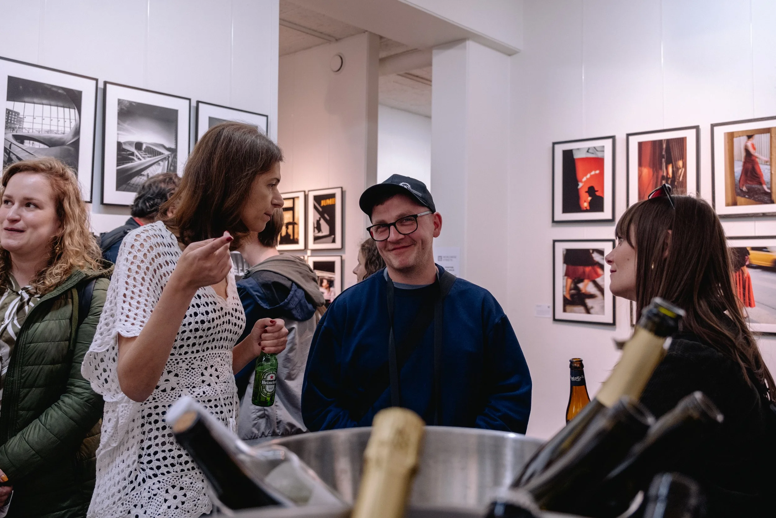People socializing at an art gallery, with framed photographs on the walls. A man with glasses and a cap is smiling, and women are engaged in conversation, holding drinks.