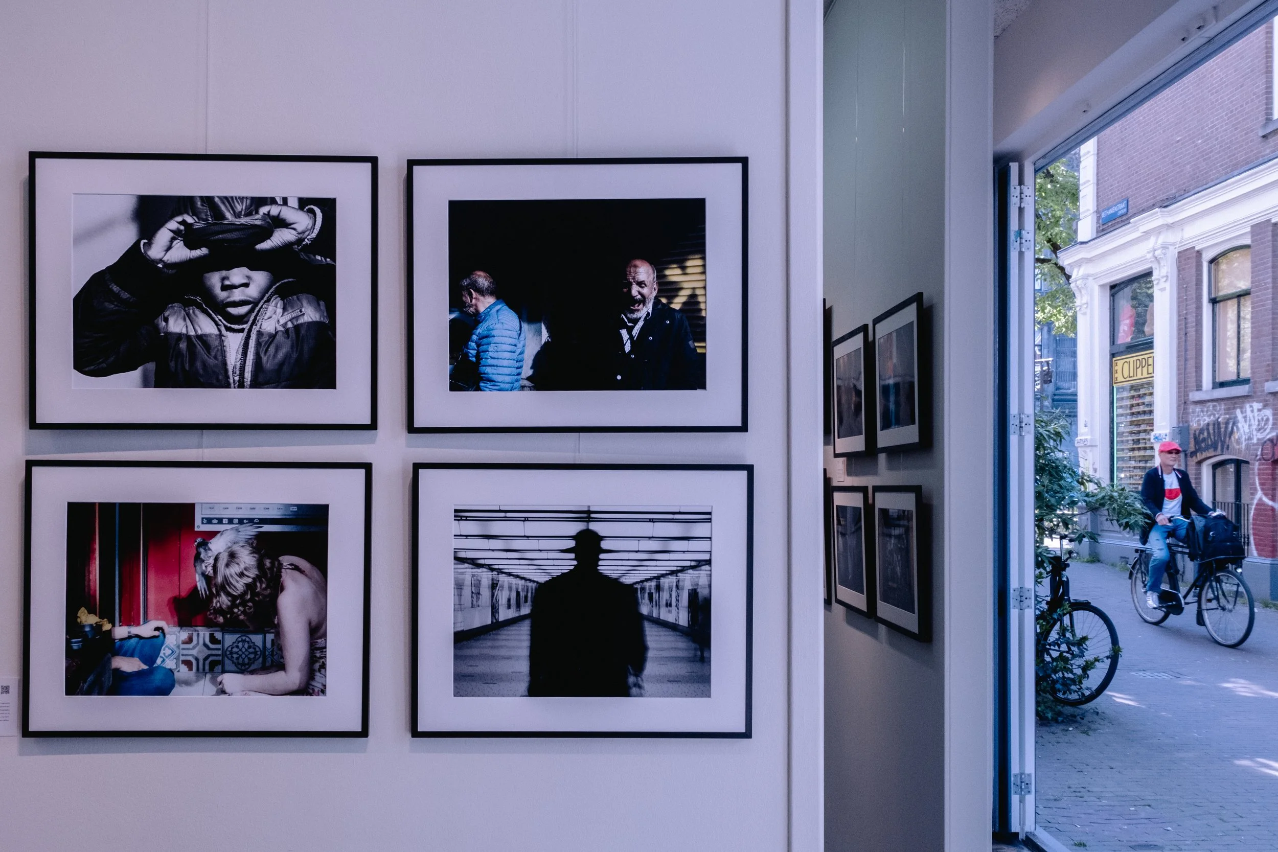 Street Photography art gallery in Amsterdam with framed photographs displayed on white walls, featuring silhouettes, reflections, and urban scenes, with a partly visible art desk and chairs in background.