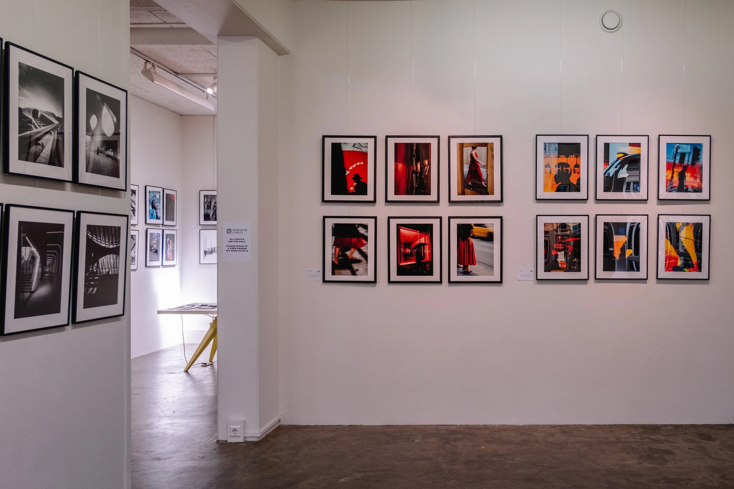 Gallery wall with framed photographs, some in black and white and others in vibrant colors, depicting urban scenes, people, and architecture in an art gallery.