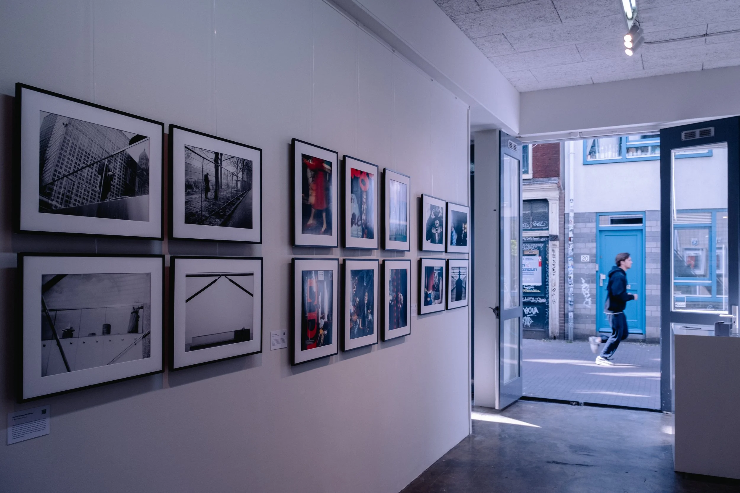 Street Photography art gallery in Amsterdam with framed photographs displayed on white walls, featuring silhouettes, reflections, and urban scenes, with a partly visible art desk and chairs in background.