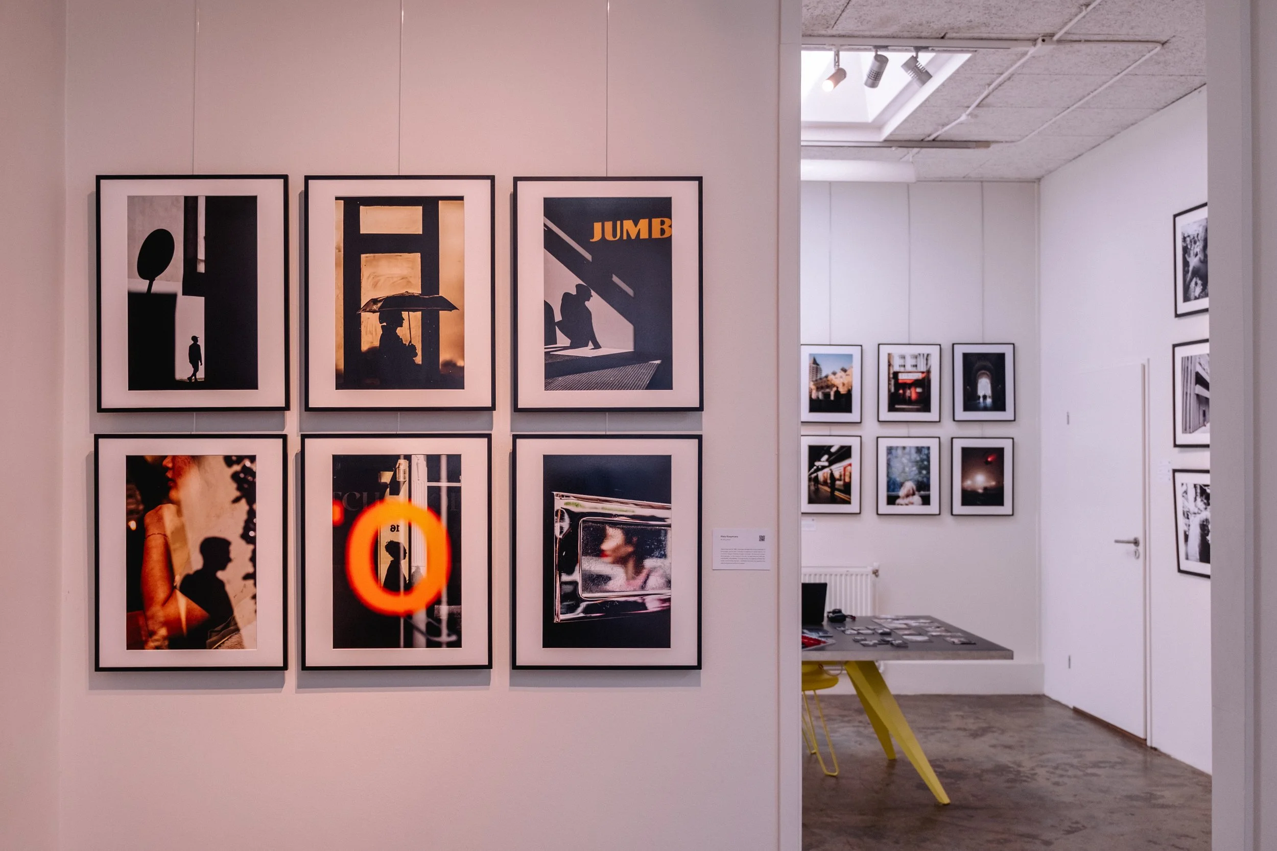 Street Photography art gallery in Amsterdam with framed photographs displayed on white walls, featuring silhouettes, reflections, and urban scenes, with a partly visible art desk and chairs in background.