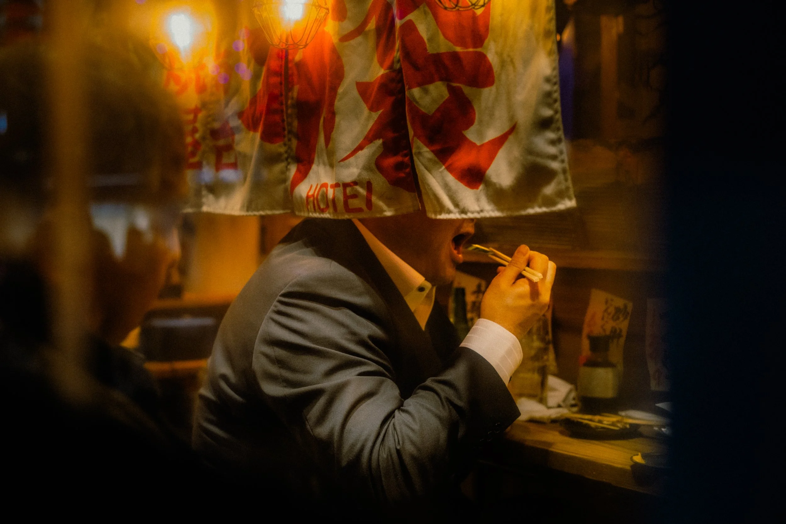 A person in a suit eating ramen noodles with chopsticks inside a dimly lit Japanese restaurant, with a curtain bearing Japanese characters and the word 'HOTEL' partially visible in the background.