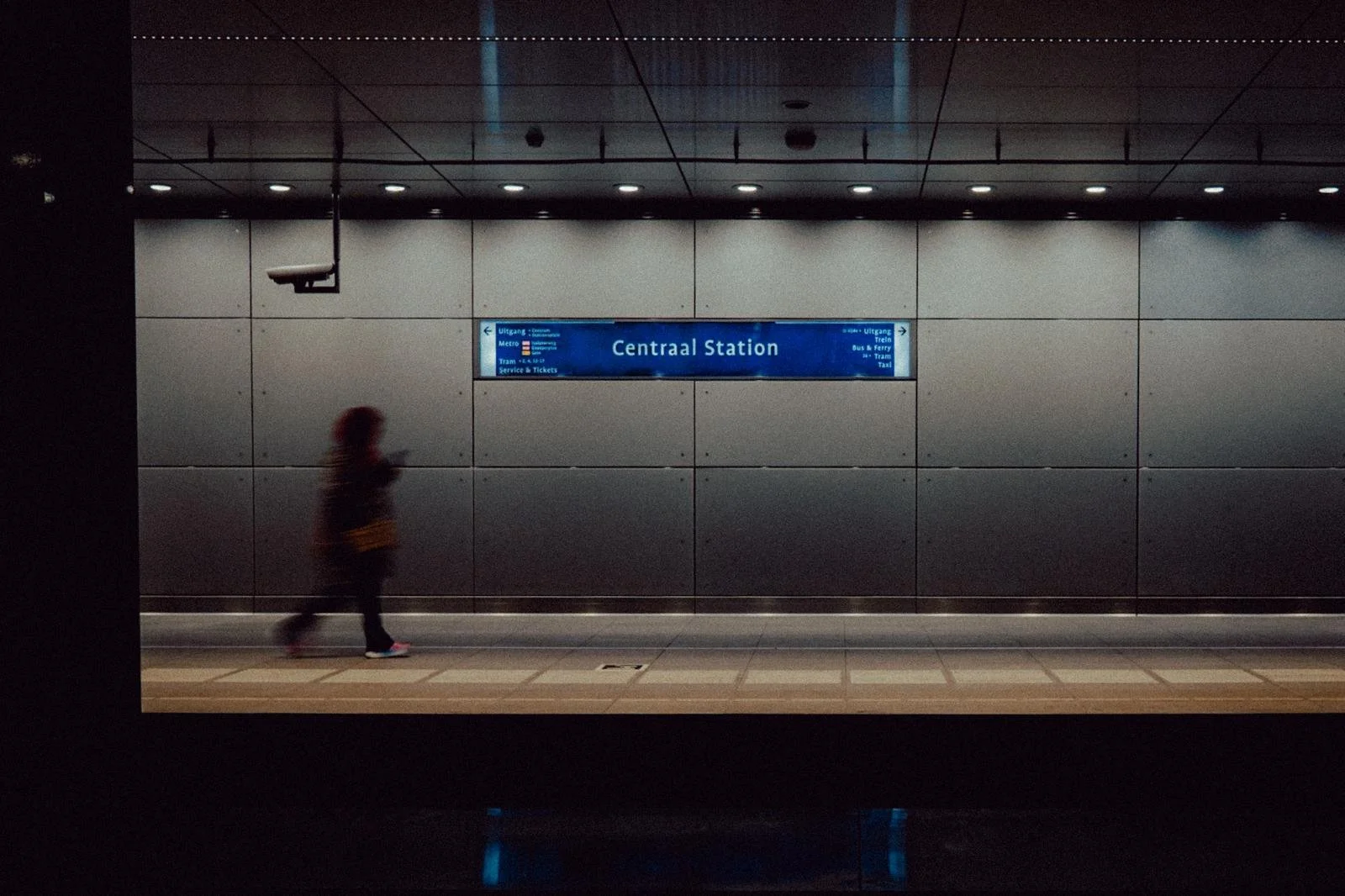A person walking in an underground metro station with a blue sign indicating 'Centraal Station' and directions for different train and tram services