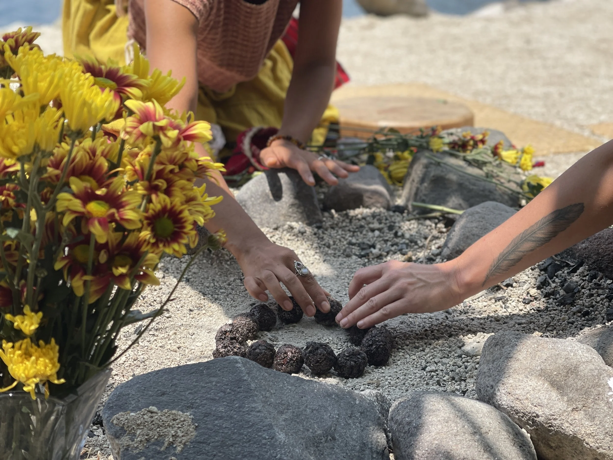 People laying stones on a memorial or grave at a beach with flowers nearby.