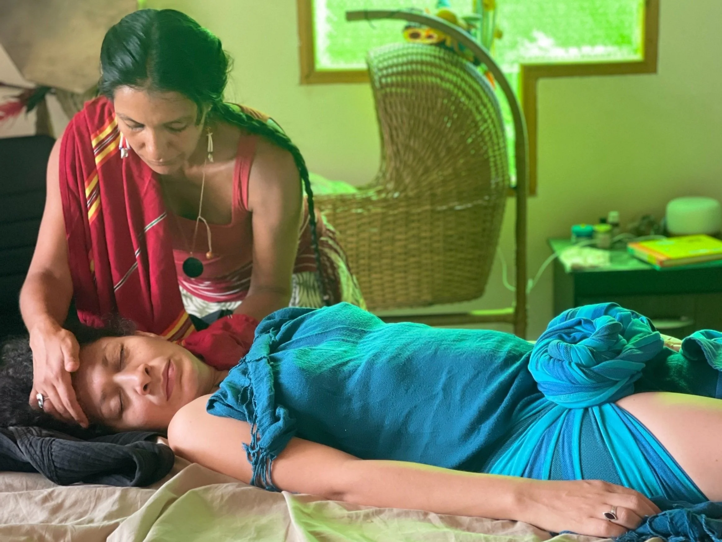 A woman with long braided hair wearing a red dress is providing massage therapy to a young woman lying on a table relaxing.