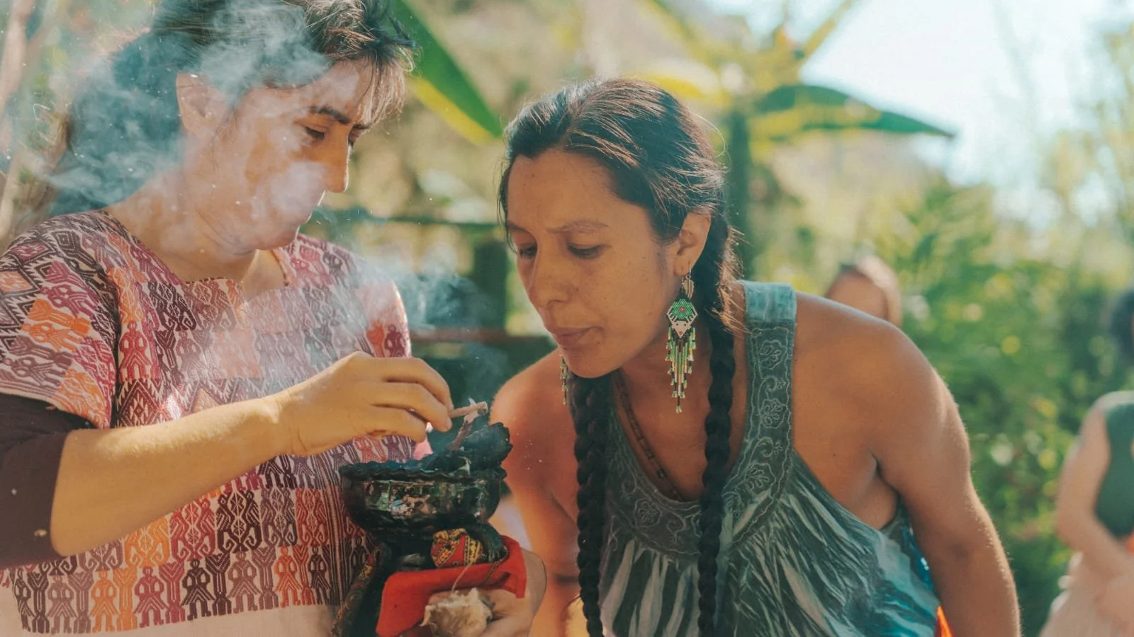 Two women engaging in a spiritual or ritualistic activity outdoors, with one woman holding an incense or ceremonial object and the other observing closely, surrounded by green foliage.
