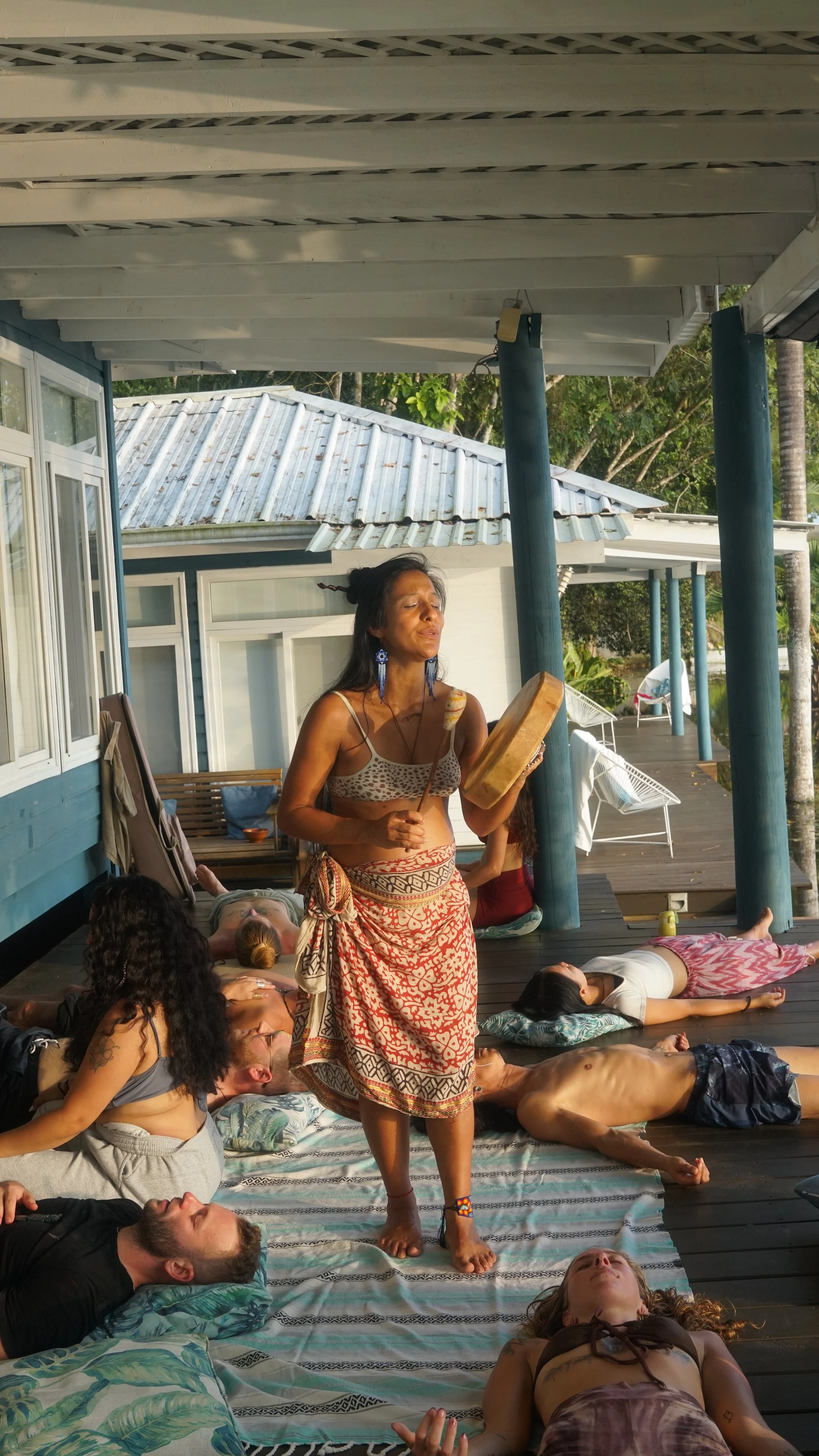 A woman standing barefoot on a wooden deck, surrounded by people lying down on mats and cushions, with some playing instruments and others relaxing, in an outdoor setting with trees and buildings in the background.