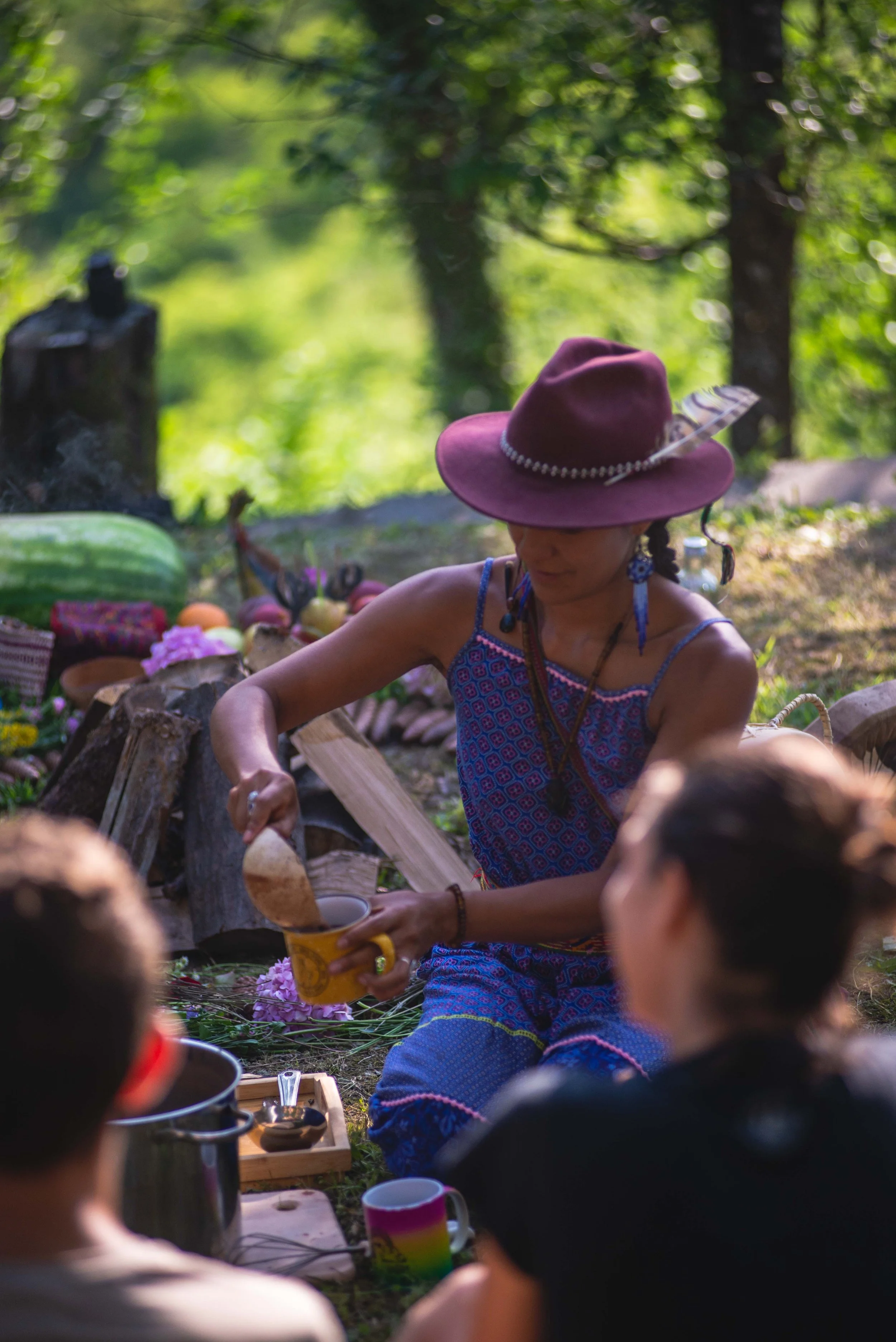 A woman wearing a purple hat with a feather and earrings is serving food in an outdoor setting with green trees in the background. There are people around her and various items, including watermelons, flowers, and cups, on the ground.