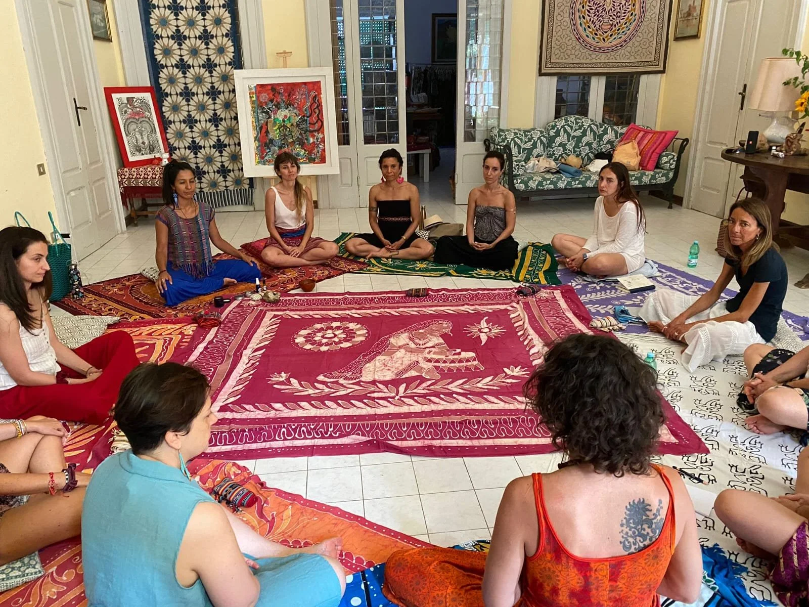 A group of women sitting in a circle on colorful rugs inside a living room, participating in a meditation or spiritual session.