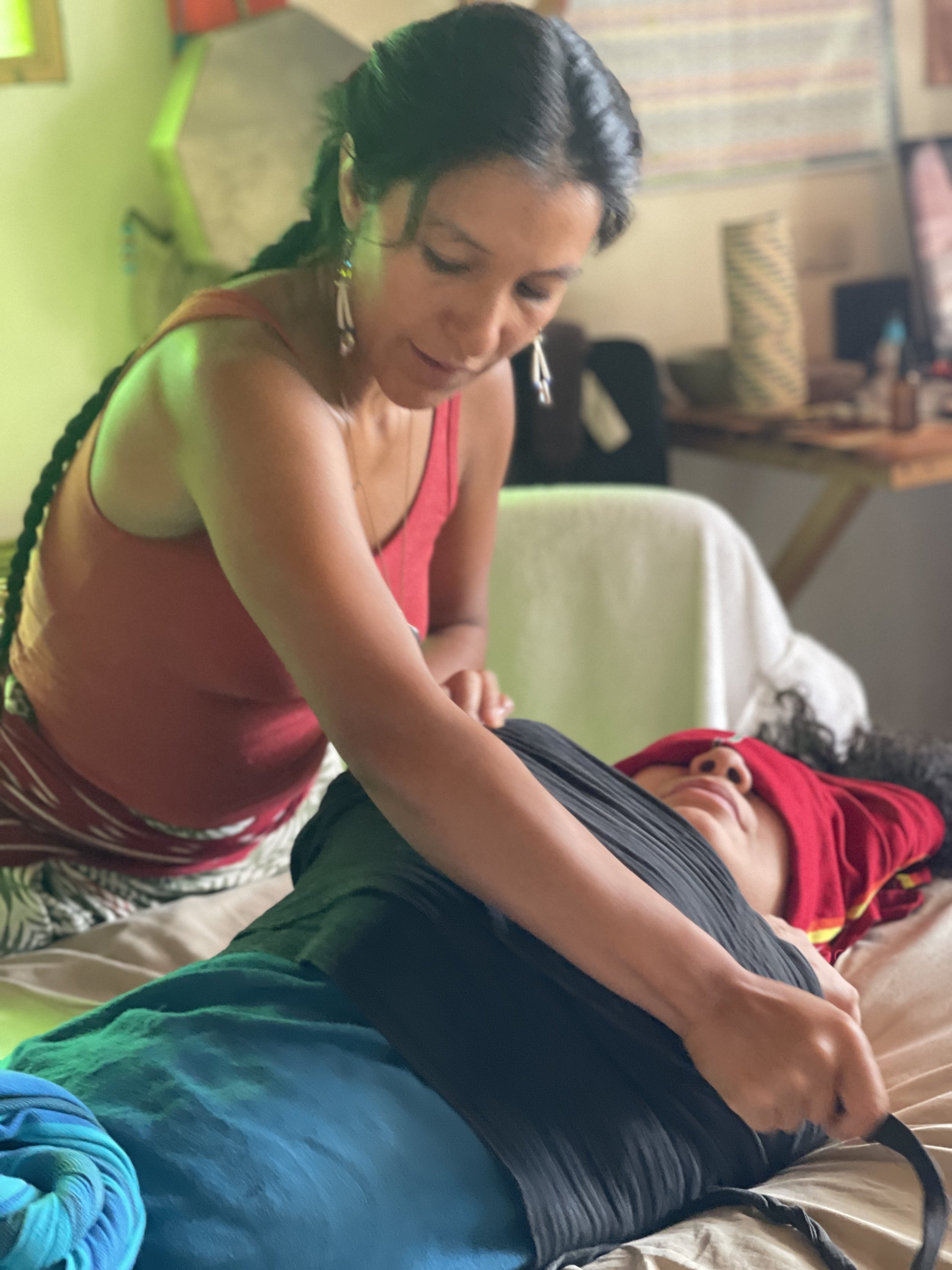 A woman is offering a massage on a woman lying down on a bed in a room with colorful decorations and a framed picture on the wall.