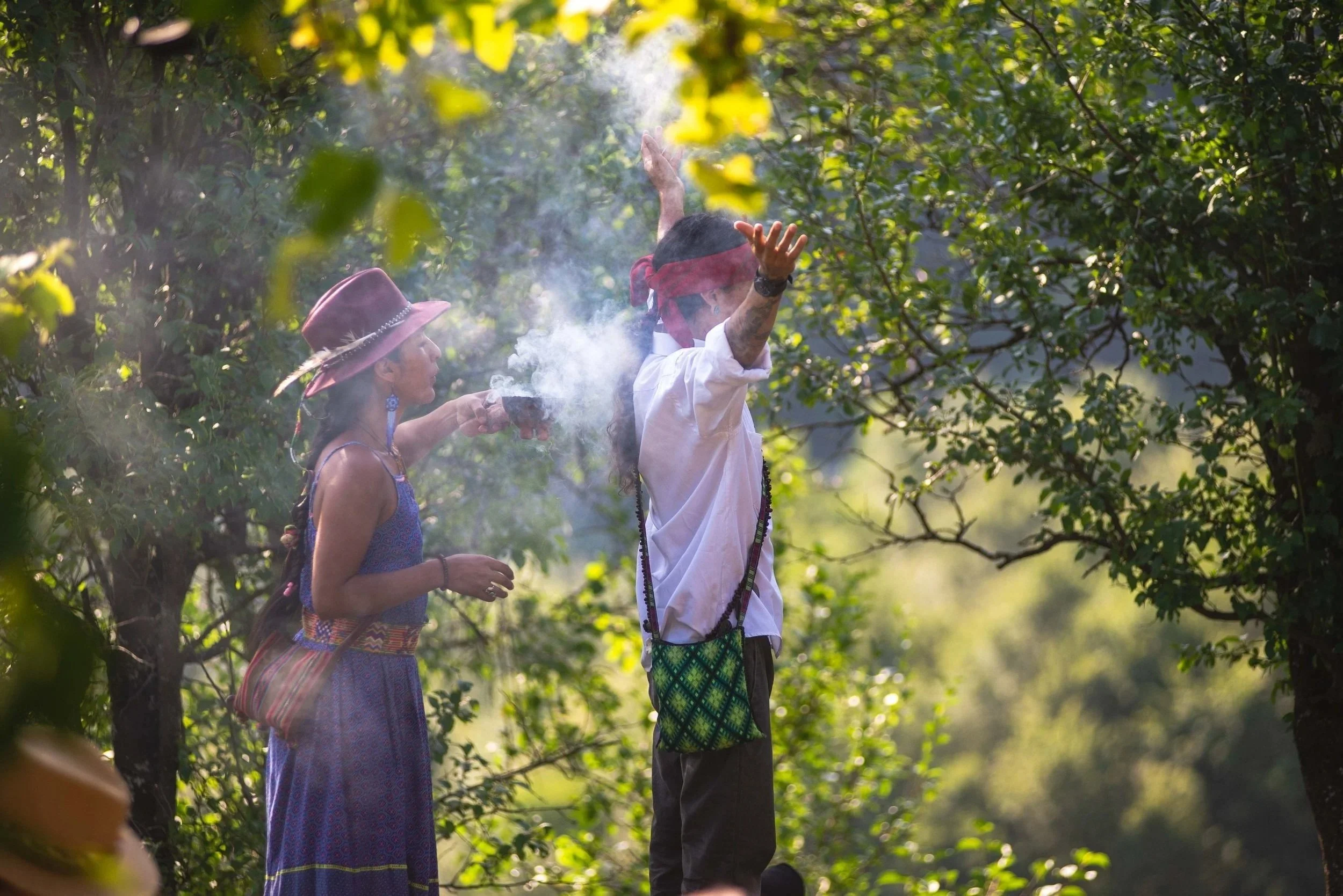 Two indigenous people, a man and a woman, standing among trees, with the man raising his hands and the woman holding an incense holder with smoke rising, in a lush green forest during daytime.