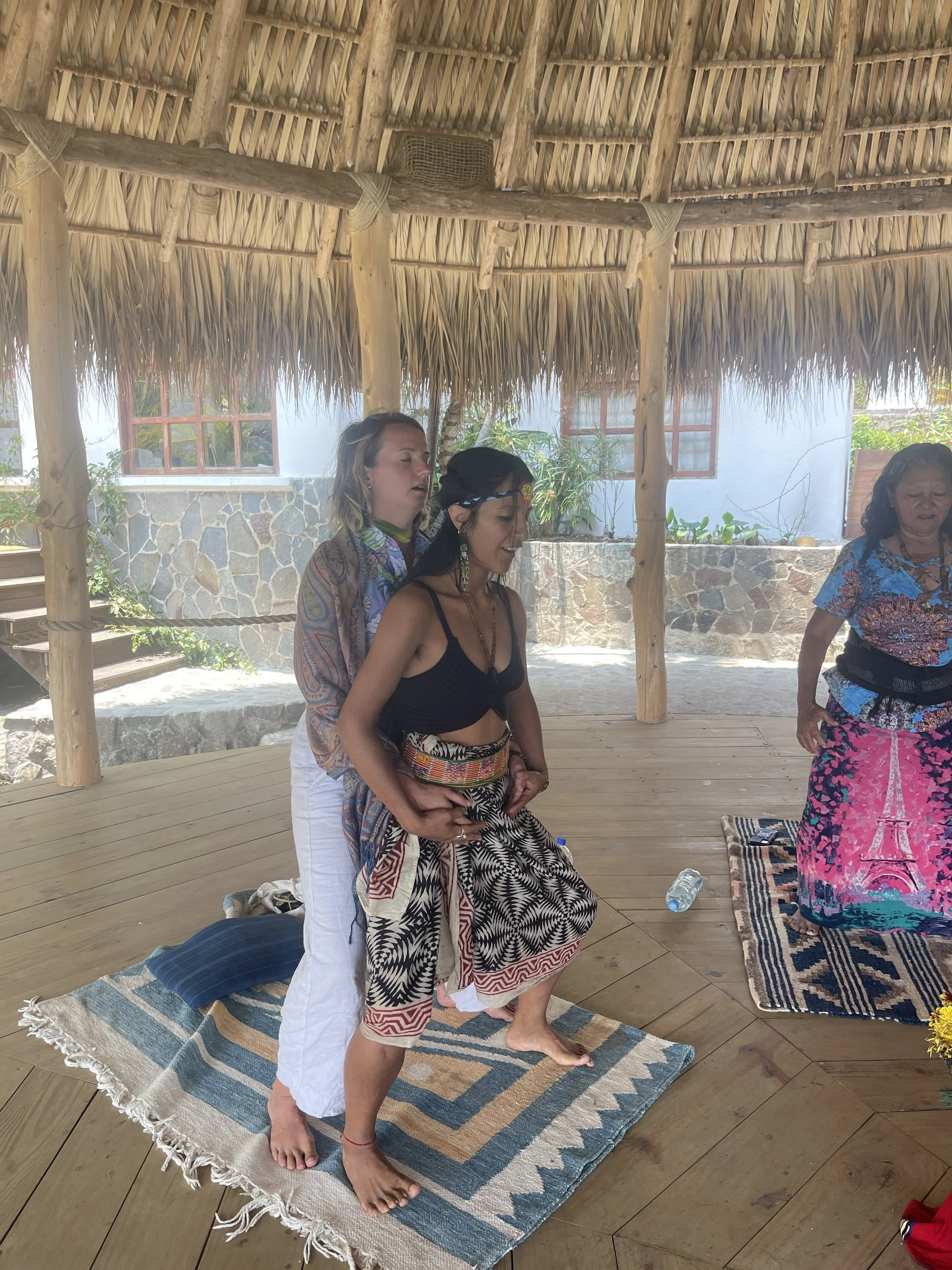 Three women practicing a spiritual or meditation exercise under a thatched roof. One woman is standing behind another woman who is seated, both with closed eyes. The third woman is standing nearby. They are on a wooden platform with mats and small rugs, with tropical plants and windows in the background.