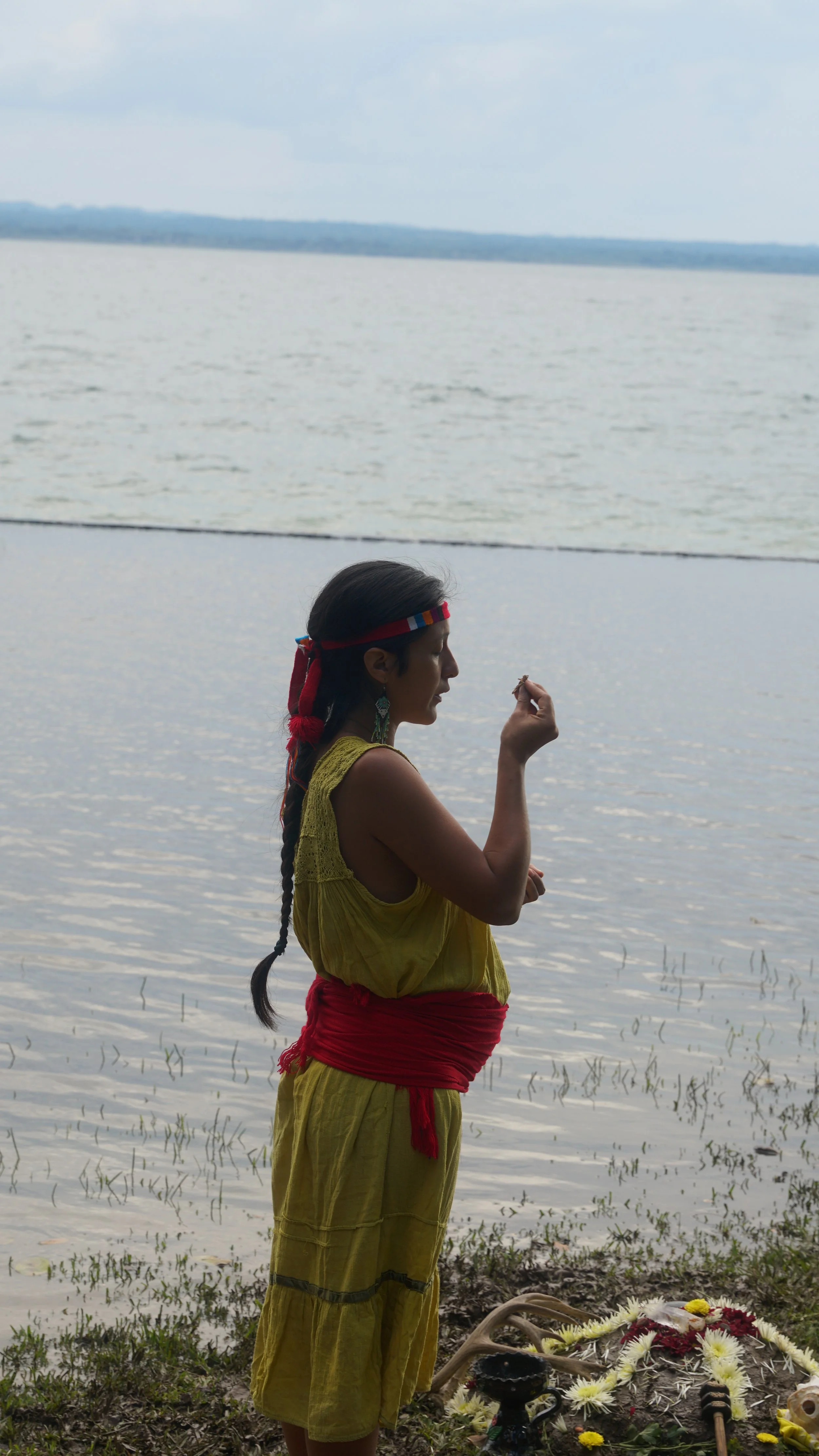 A woman dressed in traditional yellow clothing with a red sash and headband, is standing by a body of water, participating in a ritual or prayer, with flowers and offerings on the ground nearby.