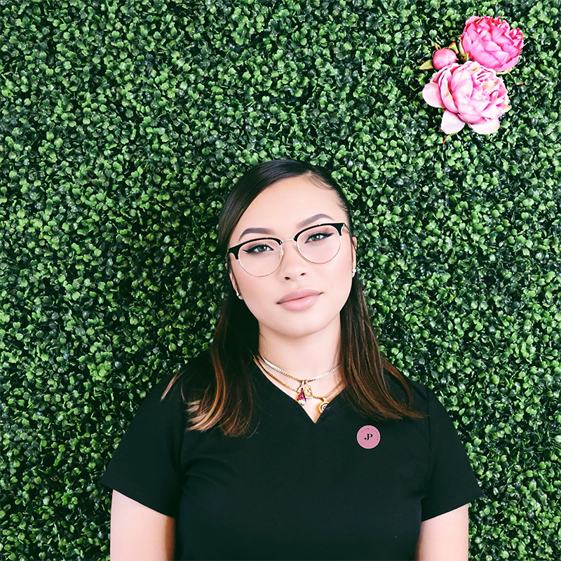 A woman with red dreadlocks smiles in front of a green leafy wall accented with pink peony flowers, wearing a black top from PINK CAVIAR Lash & Brow Spa.