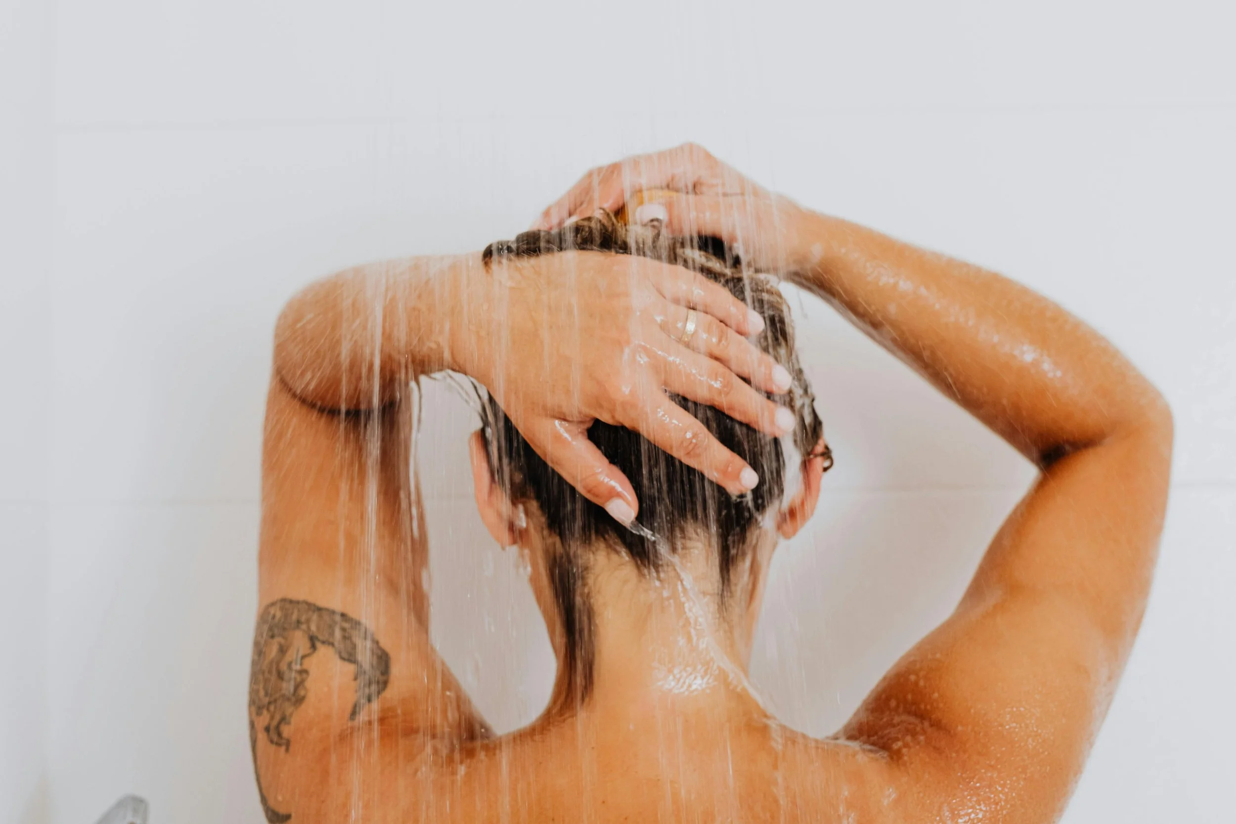 A woman with tattoos on her arm, taking a shower, with water cascading down the back of her head while she holds her hair up