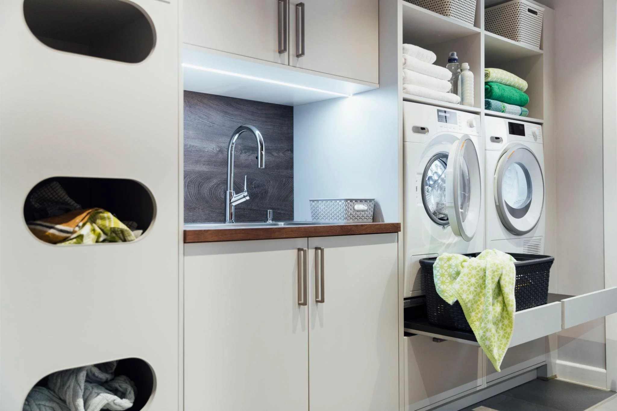 A laundry room with white cabinets, a sink, and a stack of colorful towels and linens. There are two front-loading washing machines and a black laundry basket with a green patterned towel draped over the edge.