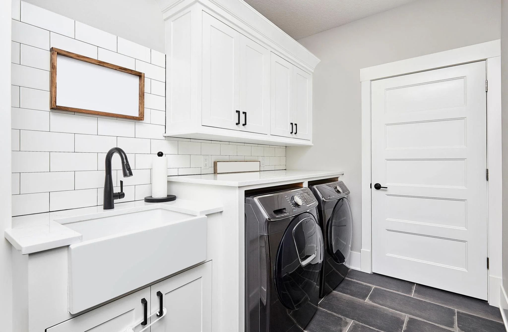 A laundry room with white cabinets, a white countertop with a black sink and faucet, white tile backsplash, and two front-loading black washer and dryer machines.