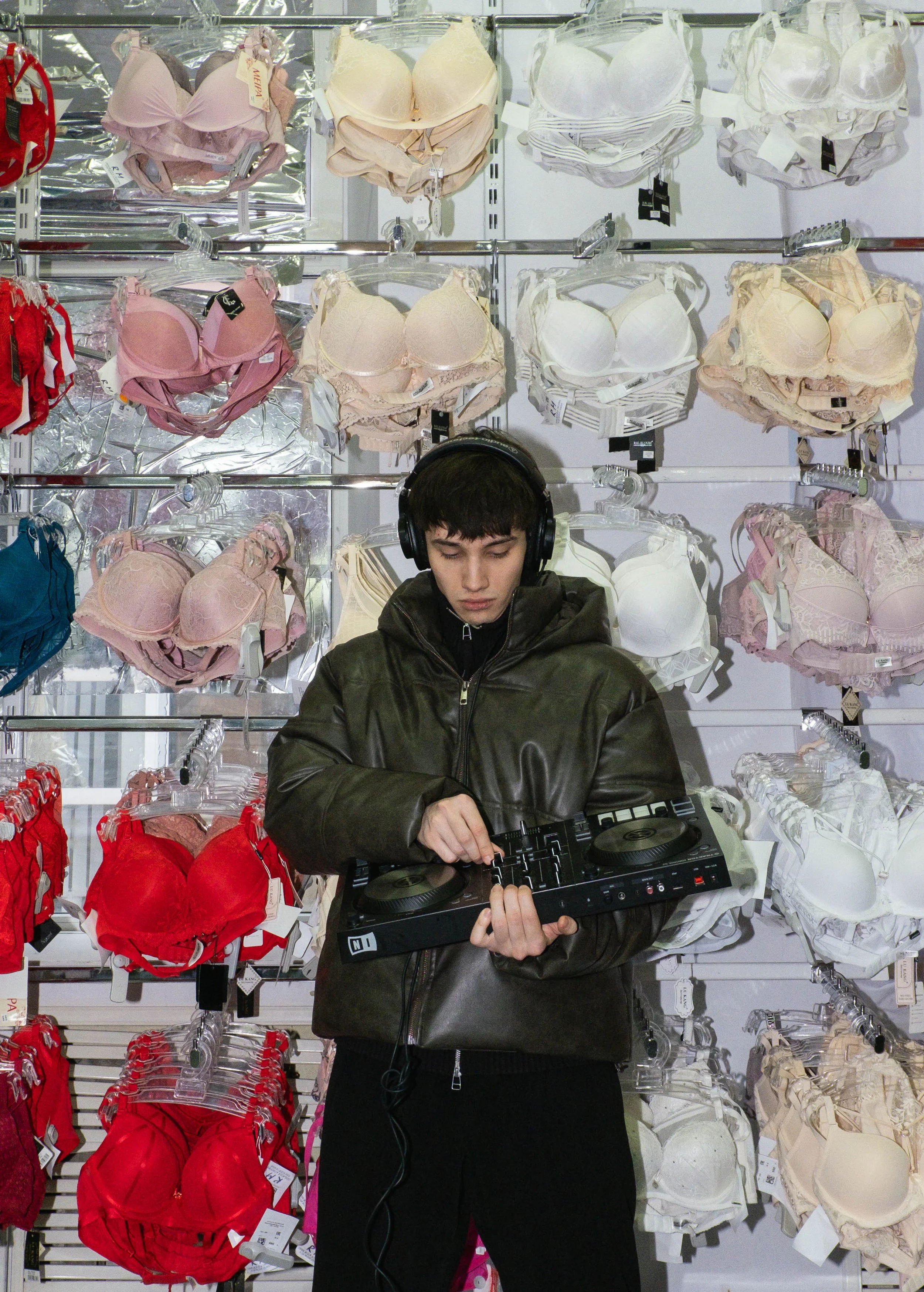 Person wearing a jacket and headphones, using DJ equipment in front of a display of bras in various colors and styles in a store.