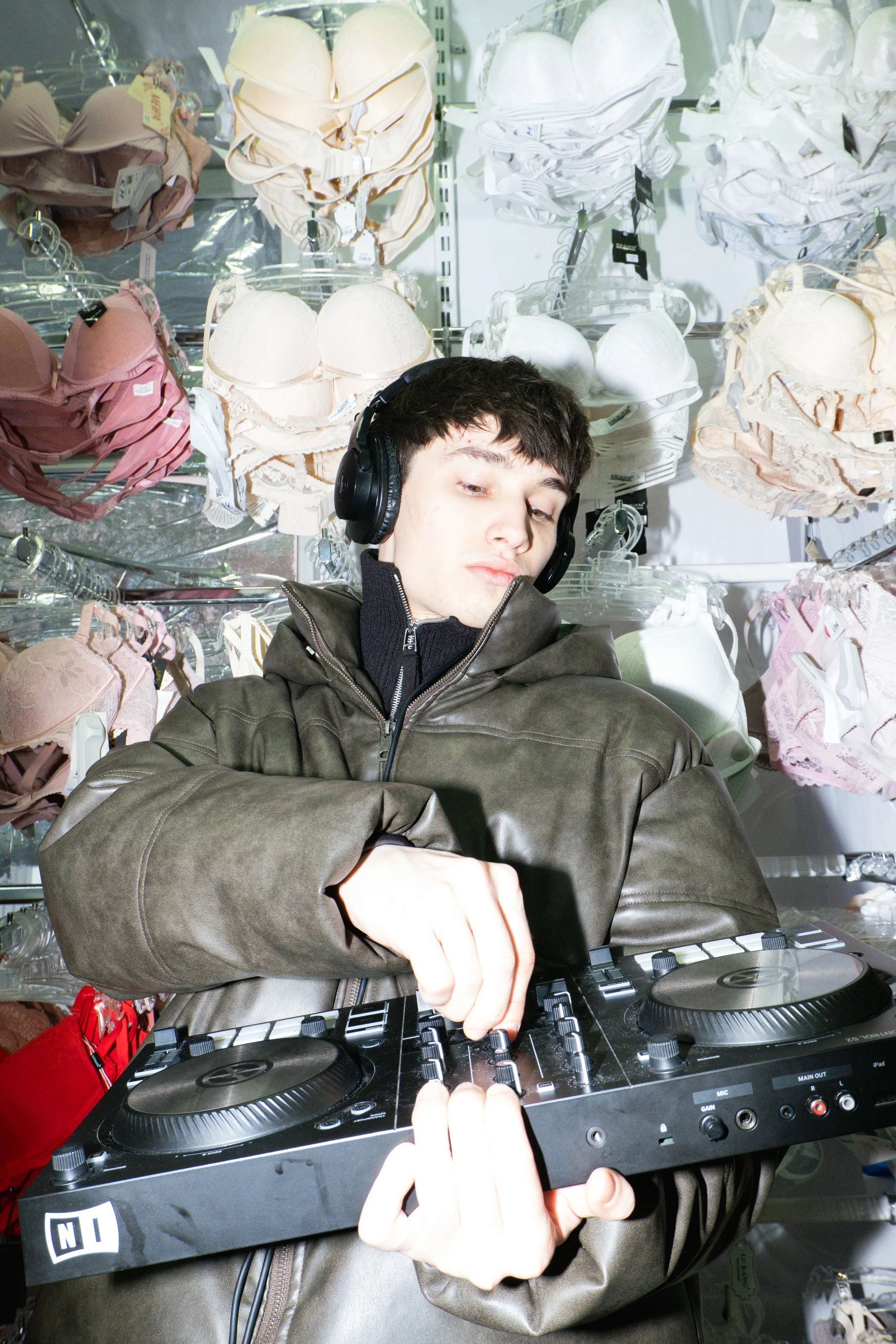 DJ wearing headphones and adjusting a mixing console in a clothing store, surrounded by bras on display.