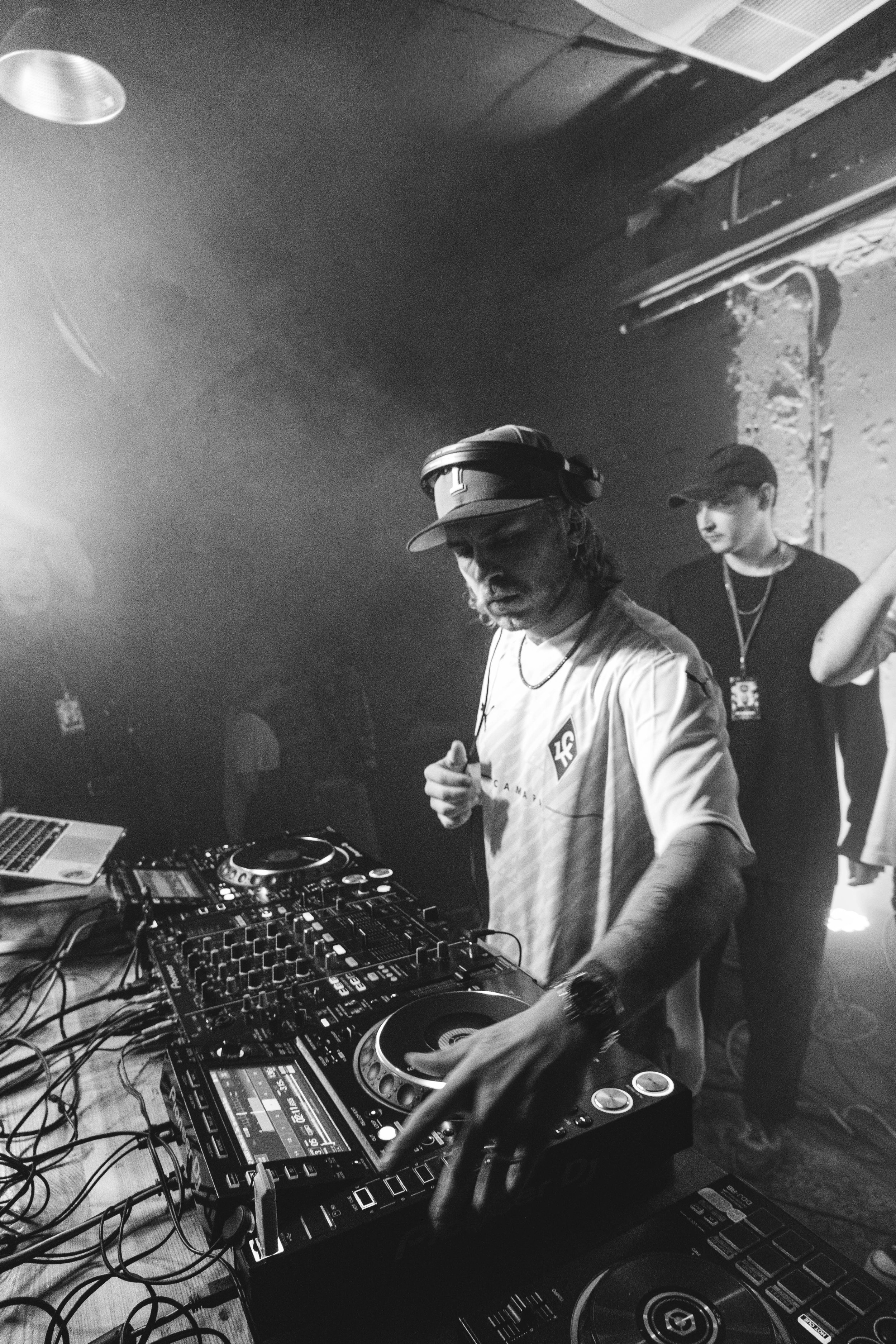 Black and white image of a DJ wearing headphones and a cap, performing at a music event. The DJ is surrounded by music equipment, including a laptop and mixers. Another person stands in the background.