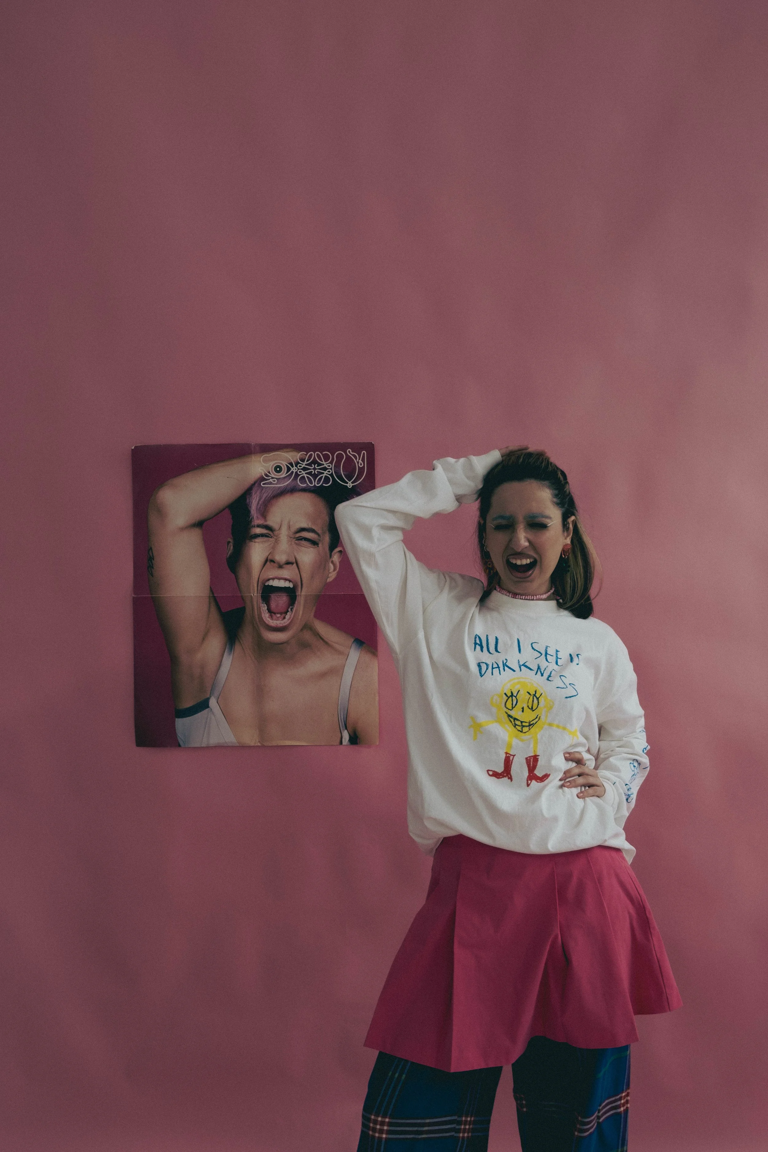 A person stands in front of a pink background, holding their head with one hand. They are wearing a white long-sleeve shirt with graphics and the text 'All I See is Darkness,' a pink skirt, and plaid pants. Beside them is a poster featuring a person